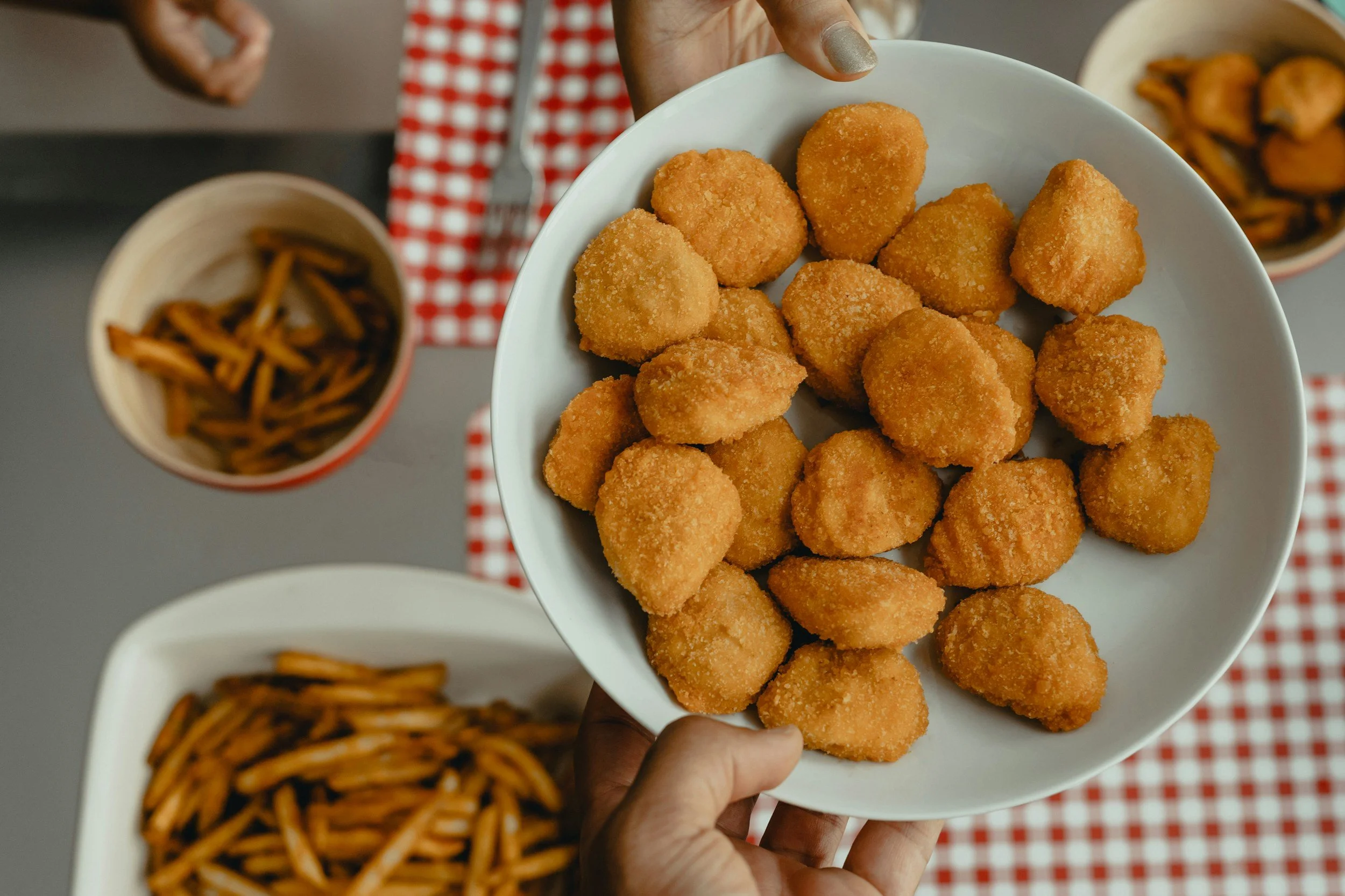 Person holding a white bowl filled with fried chicken nuggets. In the background, there's a bowl of curly fries and a cup of dipping sauce on a red checkered napkin.