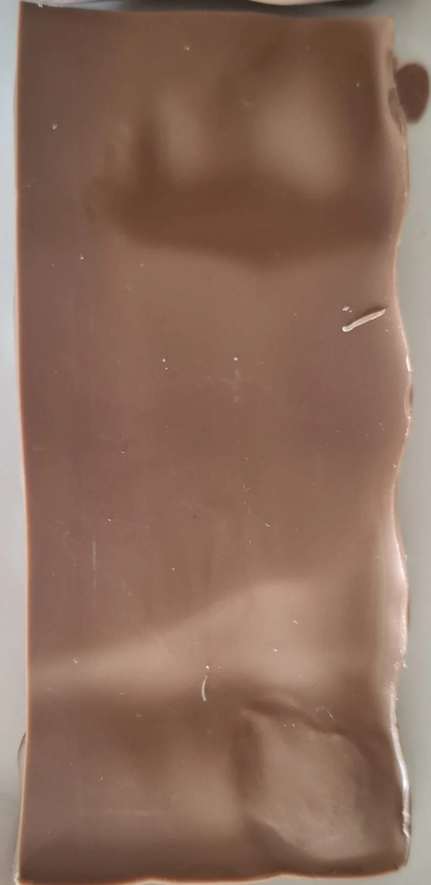 A close-up image of a piece of milk chocolate with some imperfections and a small plastic piece on the right edge.