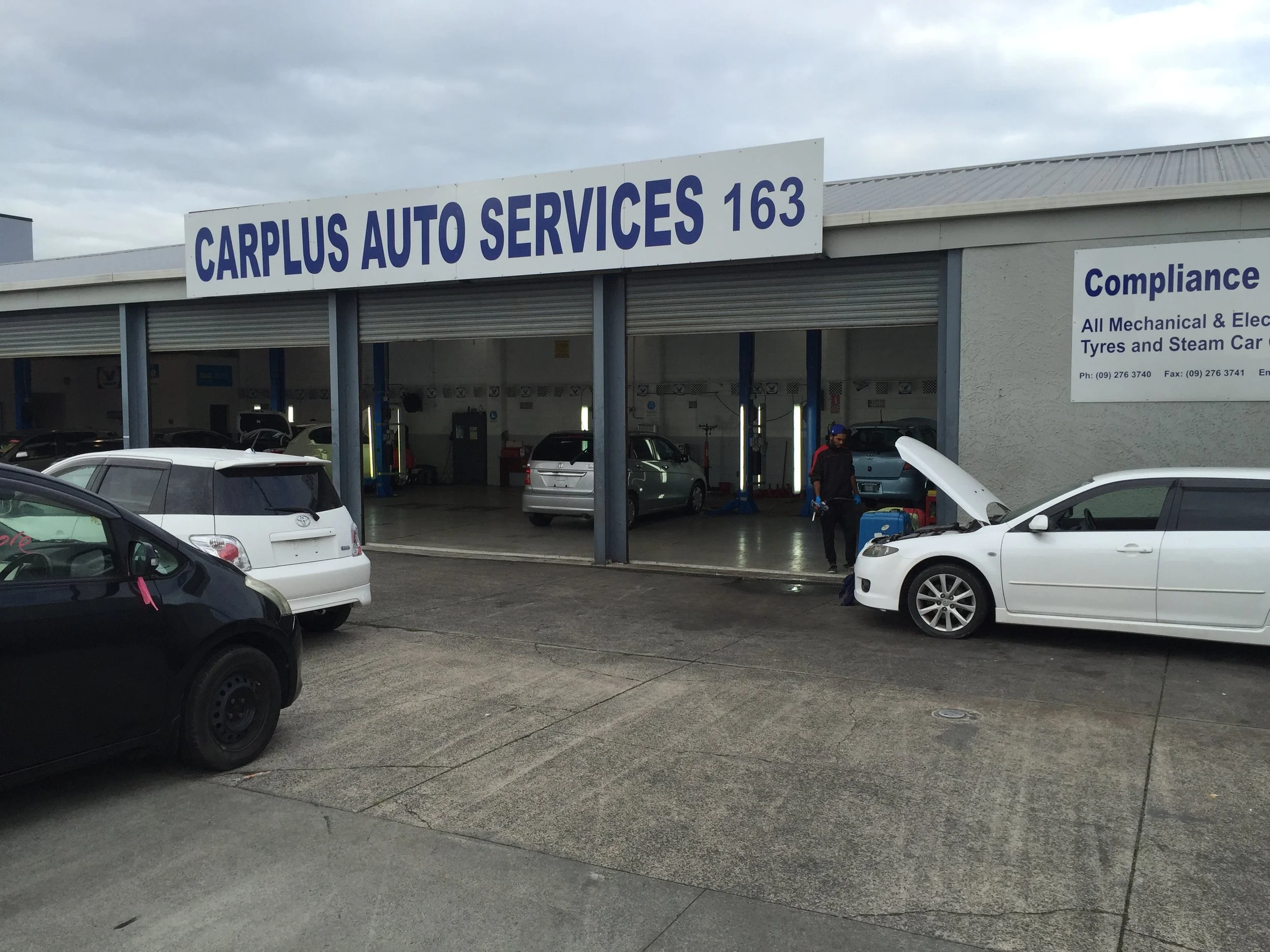 Auto repair shop named Carplus Auto Services with open garage bays, several cars inside and outside, and a worker inspecting a white car with its hood open.