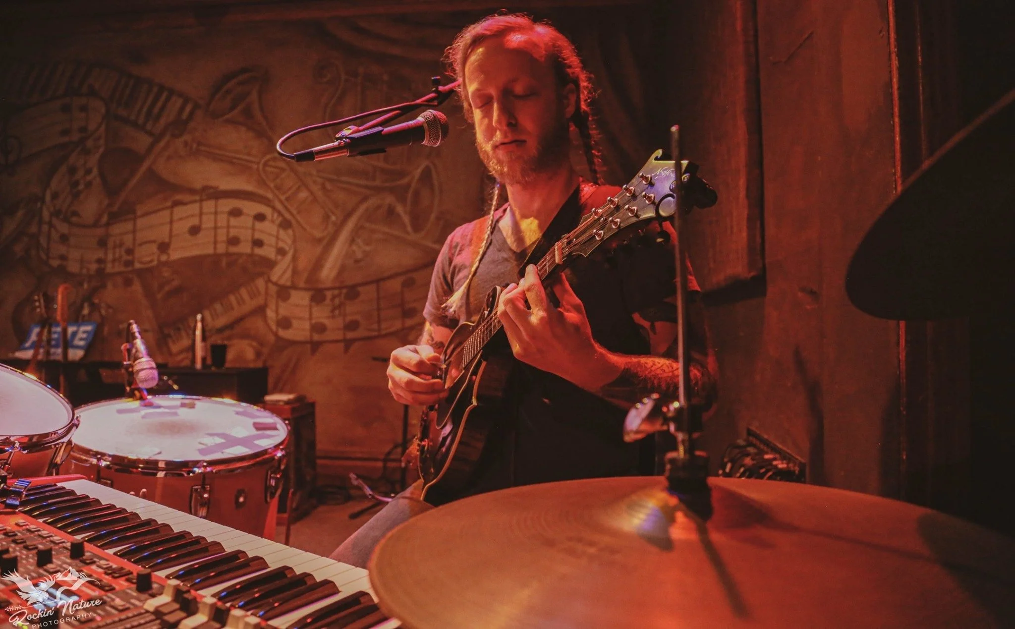 Musician with braids playing a mandolin and drums in a dimly lit venue with a painted musical notes mural on the wall behind him.