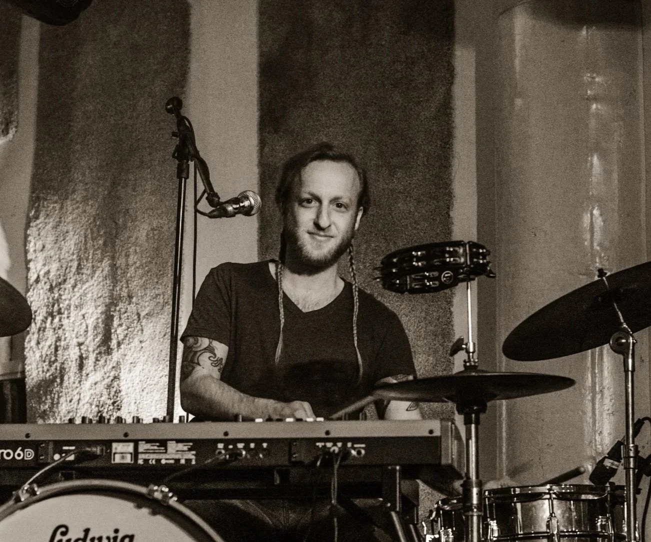 A man with a beard and tattoos is sitting behind a drum set and a keyboard, smiling at the camera, on stage at a live performance.