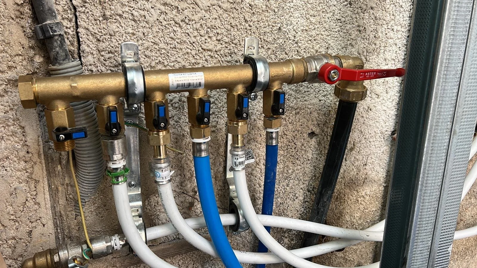 Installation de plomberie avec tuyaux en cuivre, vannes à levier rouge et commande noire, fixée contre un mur en béton, avec divers raccords et manchons.