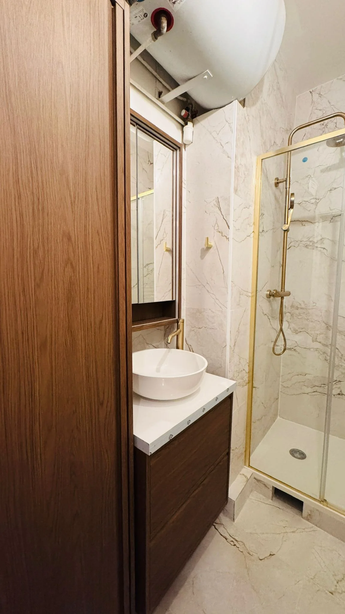 A small bathroom with marble walls and floor, a vessel sink on a wooden vanity, a mirror cabinet, and a glass shower enclosure with gold trim.