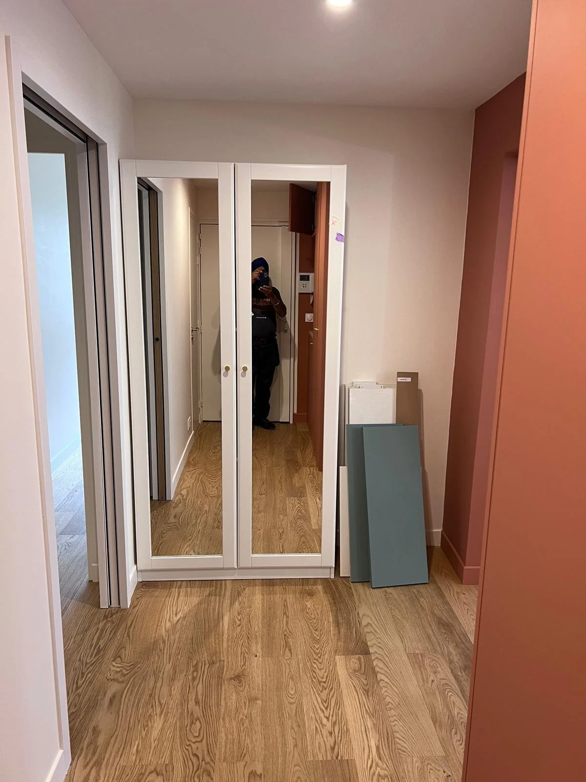 Un espace intérieur avec un grand miroir mural, des murs blanc et rose, un sol en bois clair, et des panneaux de couleur gris et blanc à droite. On peut voir une personne en arrière-plan près d'un porte-manteau ou d'une zone d'entrée.