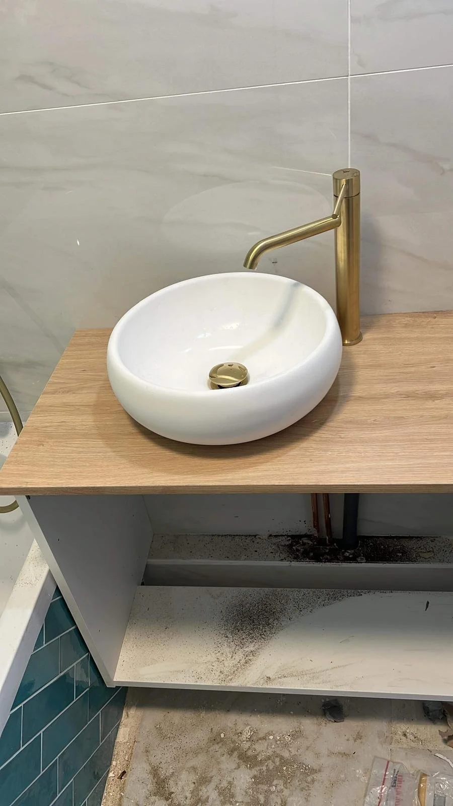 Lavabo rond blanc avec robinet en métal doré sur un meuble en bois, arrière-plan avec murs en carrelage gris. Sous le meuble, saleté ou débris au sol.