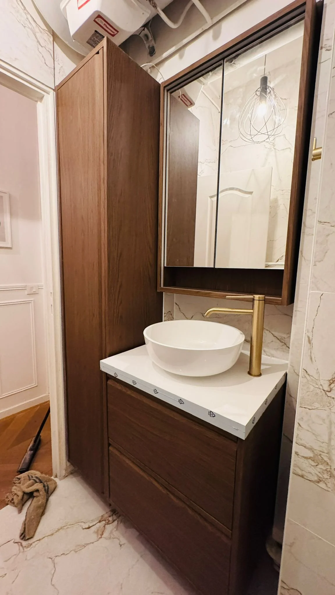 Bathroom with wooden vanity, round vessel sink, gold faucet, mirrored cabinet, and marble walls and floor.
