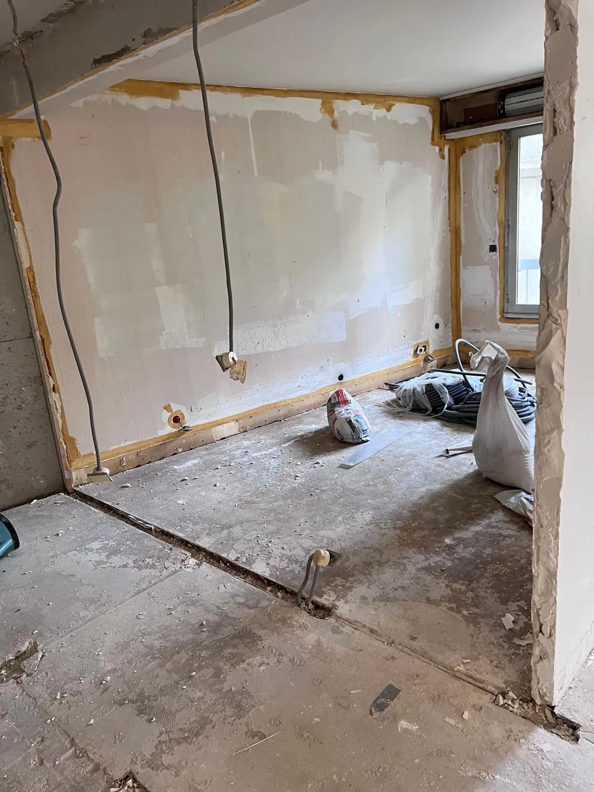 Une pièce en cours de rénovation avec des murs en cours de peinture ou de plâtrage, des câbles électriques suspendus, une fenêtre, et des outils de construction au sol.