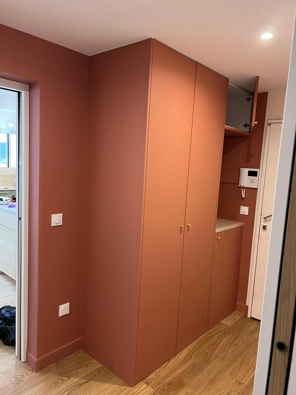 Un placard en bois dans une entrée, avec des portes fermées, une armoire ouverte et un interphone sur le mur.