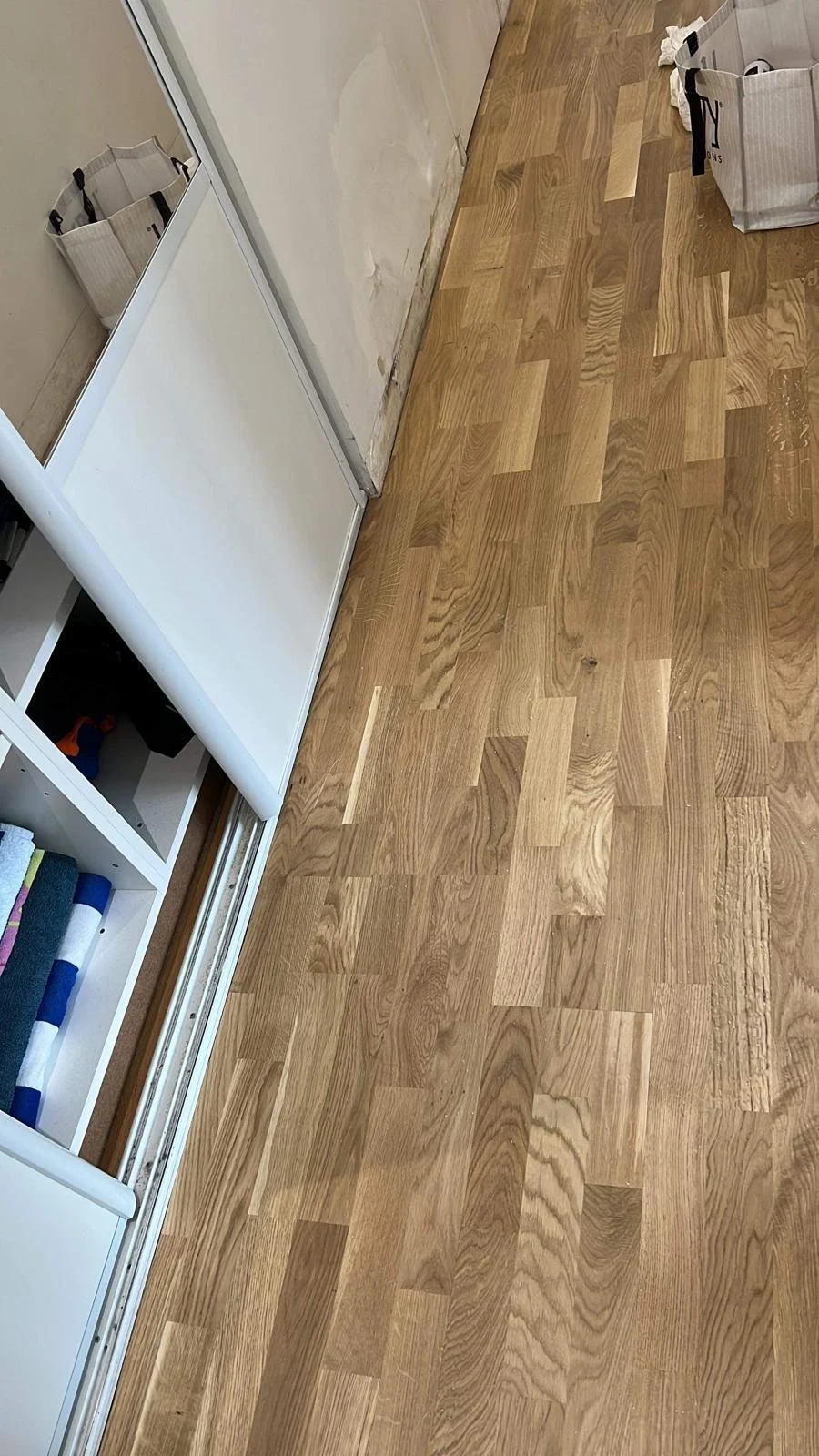 Sol en bois clair avec une étagère blanche contenant des articles de rangement, une porte en miroir, et un sac en carton dans un intérieur en cours de rénovation.