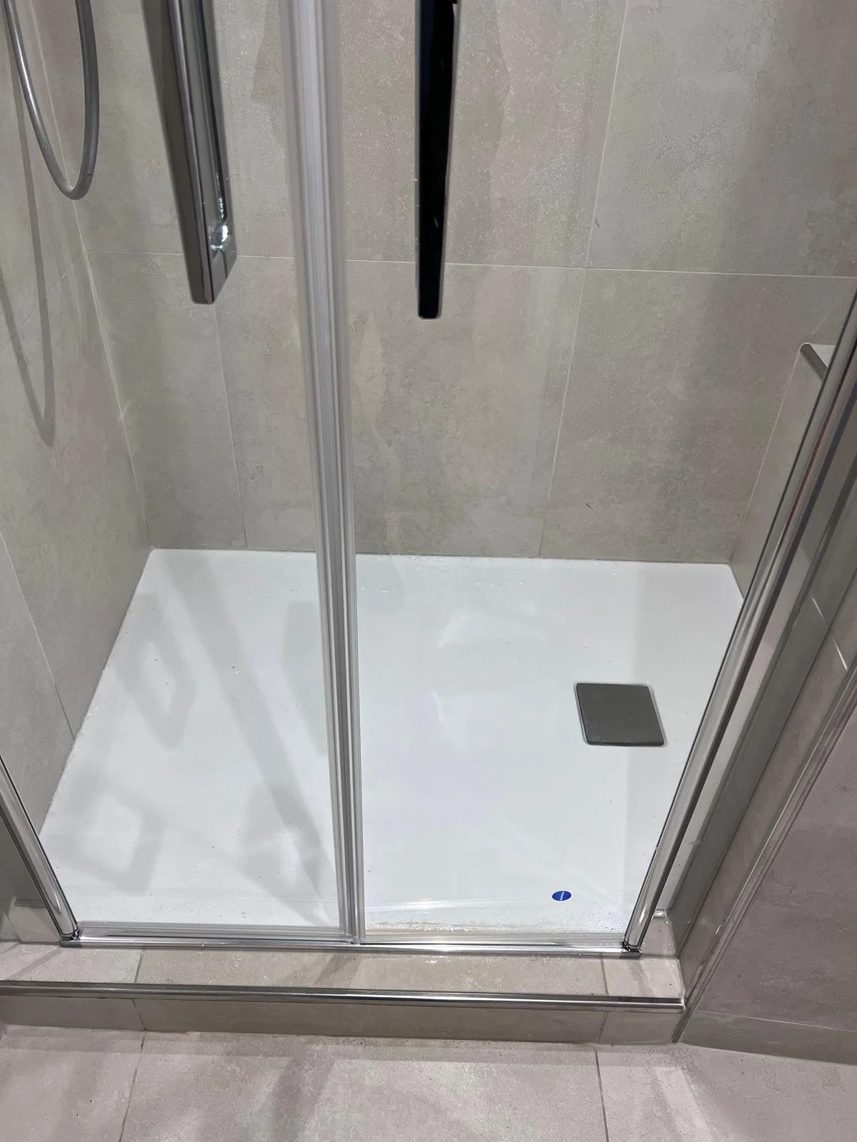 Douche à cabine en verre avec un sol blanc et un drain au coin, murs en carrelage beige.