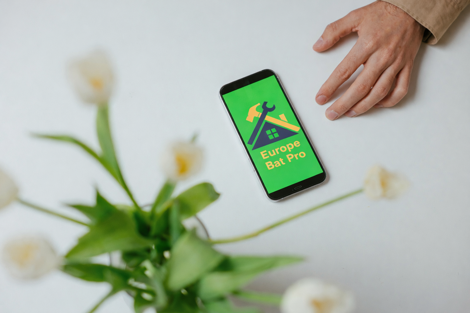 Smartphone on a white surface displaying the logo and text "Europe Bat Pro", next to a person's hand and some white flowers and green leaves.