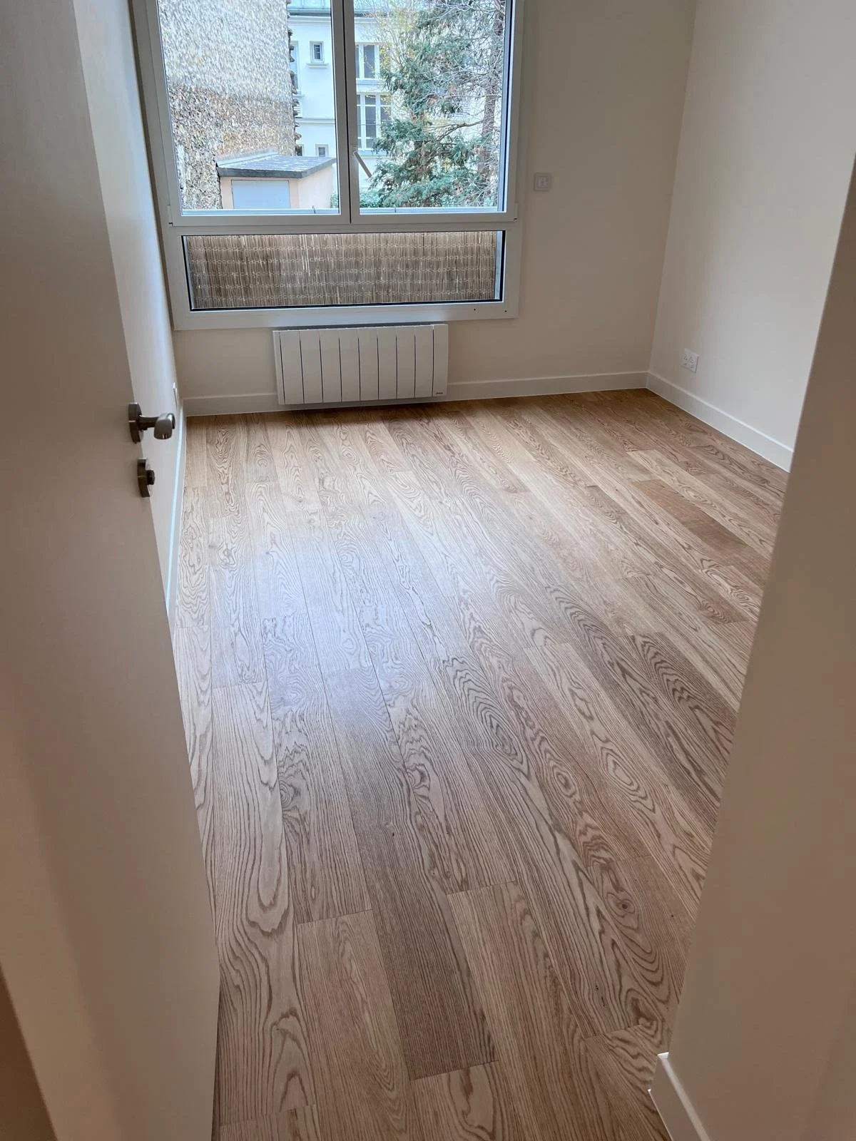 Pièce vide avec parquet en bois clair, grande fenêtre, murs blancs, chauffage électrique sous la fenêtre.