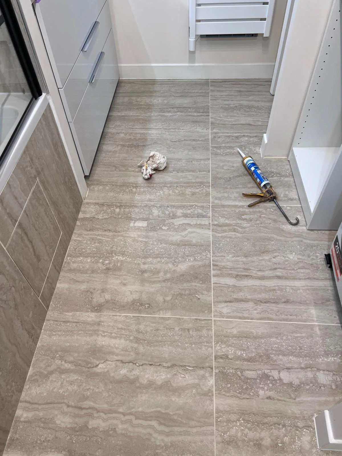Sol en carrelage beige avec des outils de réparation, notamment un tube de colle et un crochet, ainsi qu'un chiffon, dans une pièce probablement une cuisine ou une salle de bain.