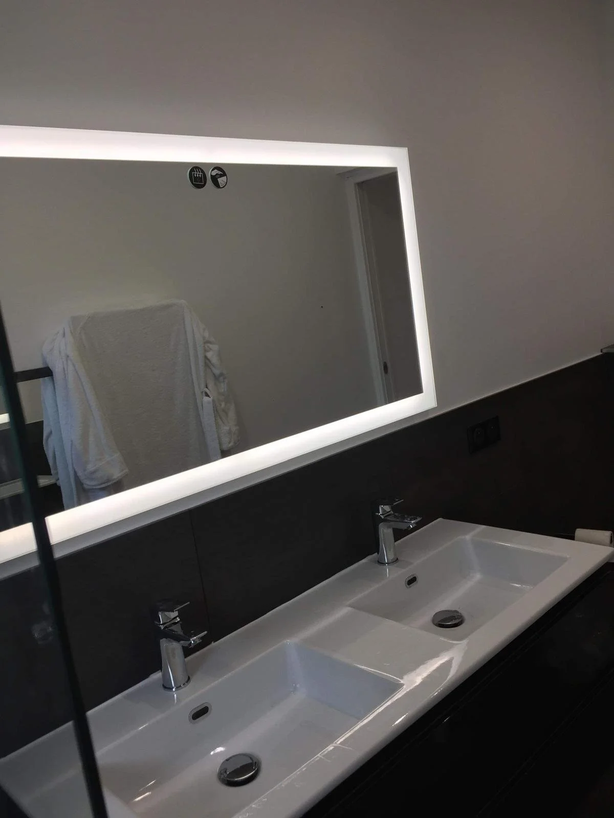 Salle de bain avec deux lavabos, un miroir avec éclairage LED, une serviette blanche suspendue, et un mur noir