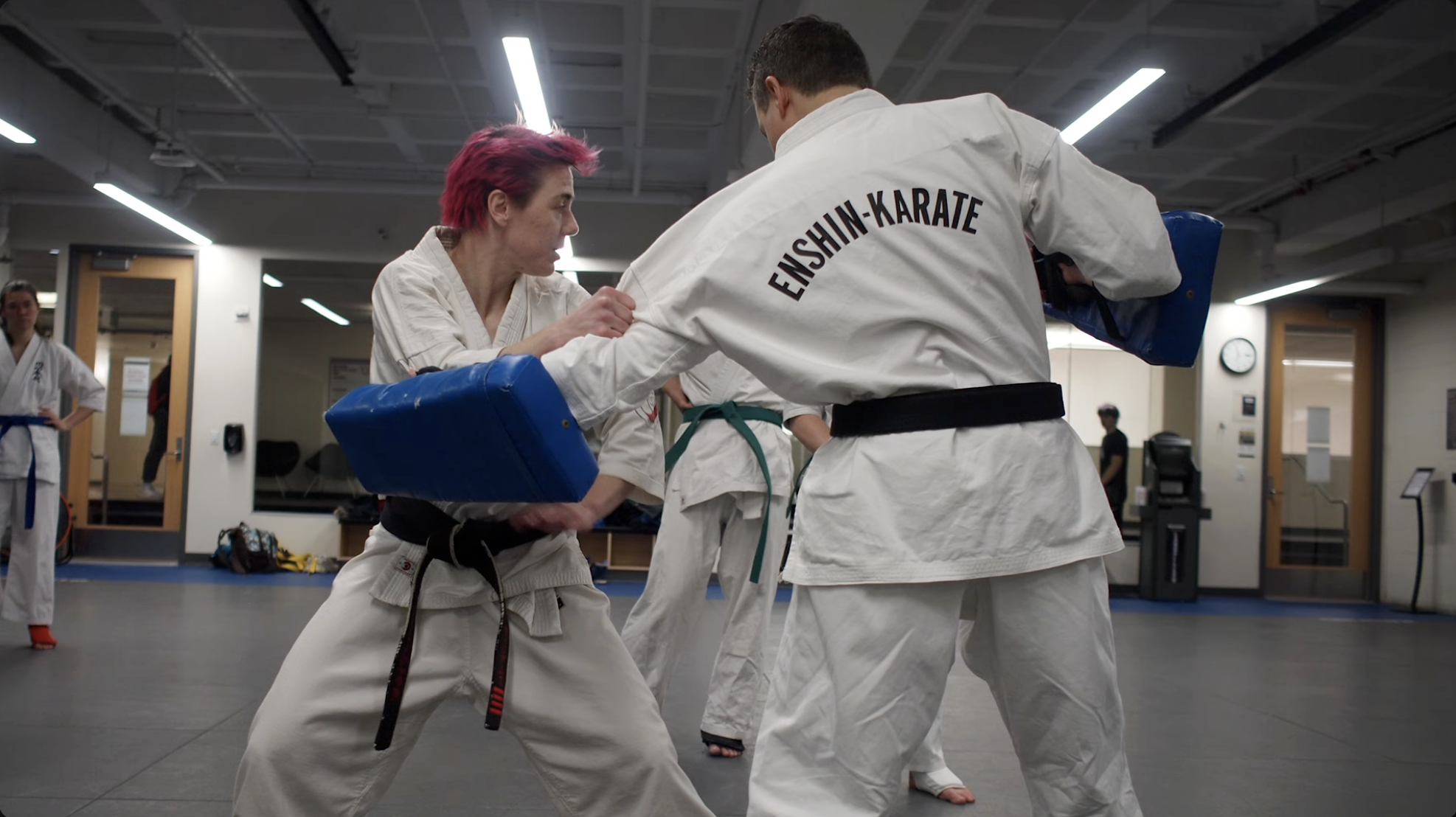 A woman with pink hair practicing martial arts with a man wearing a white gi labeled 'Enshin-Karate' in a dojo with other students in the background.