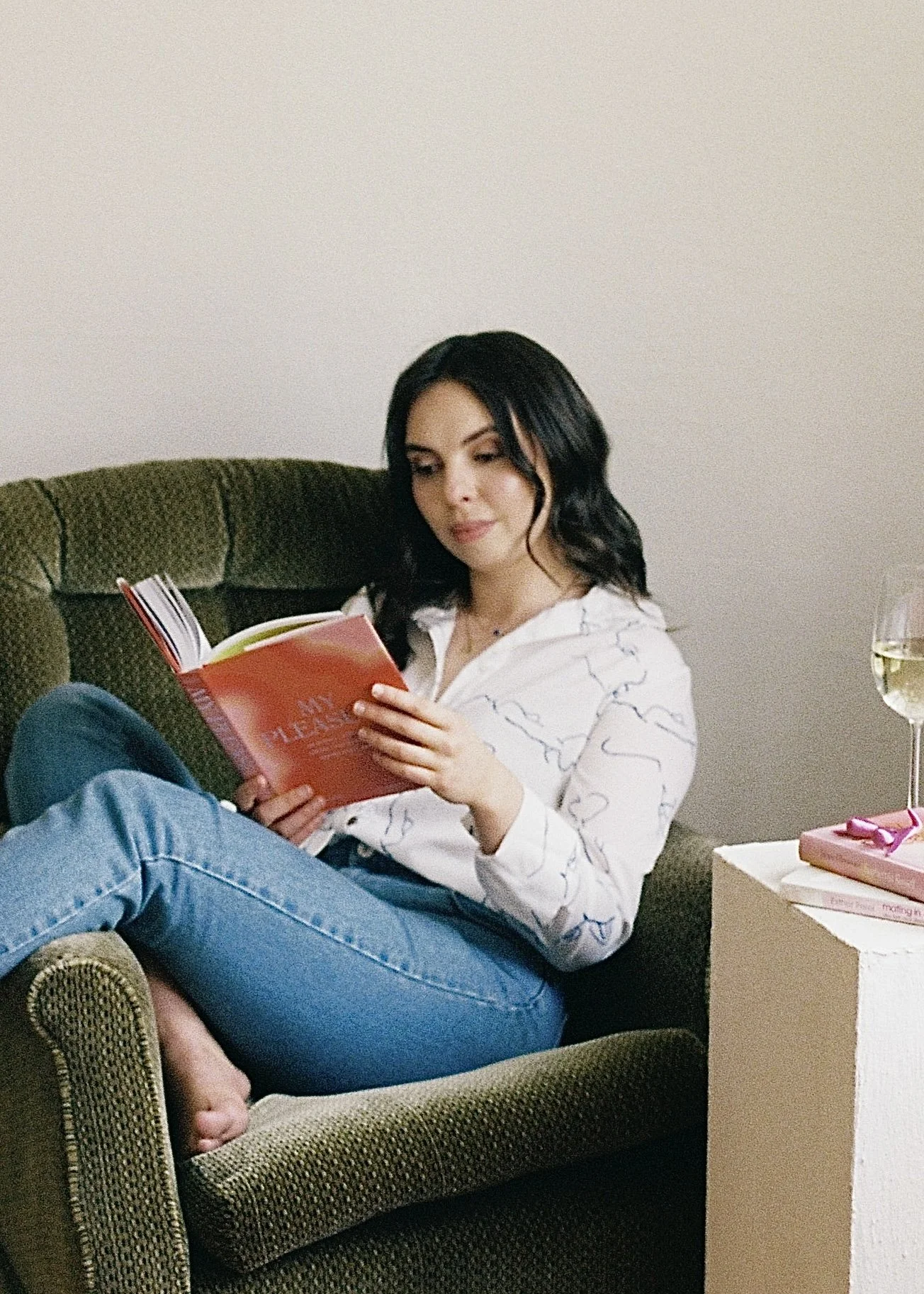 Eleni Gabrielides, a Canberra-based sexologist with brown hair, sitting in an armchair reading the book “My Pleasure,” deeply engaged in learning about sexual health and pleasure.