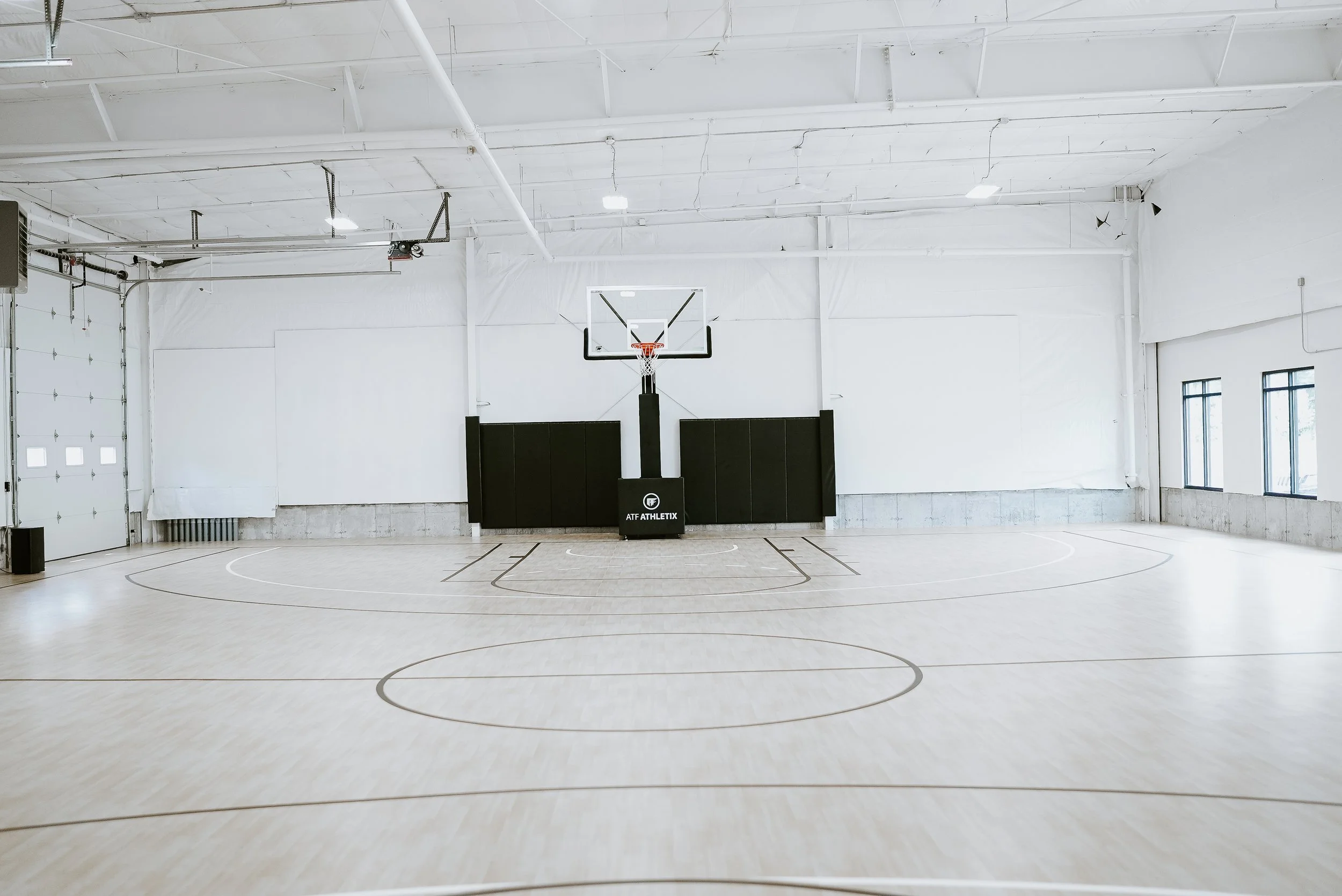 Empty indoor basketball court with a hoop, black padding, and windows along the side walls.