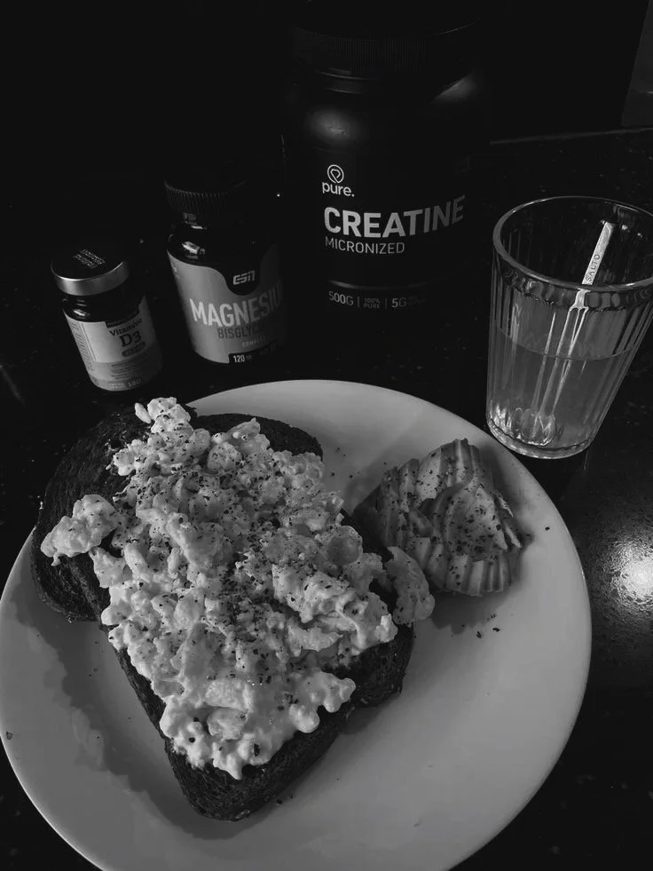 A plate with a slice of bread topped with scrambled eggs and a side of sliced fruit. Behind the plate, there are supplements and a container of creatine powder, with a glass of water on the right.