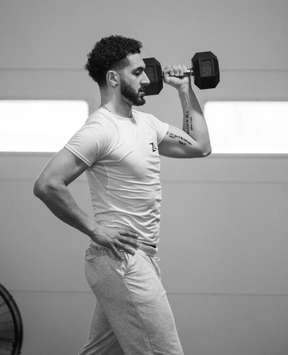 A man in a fitness center lifting a dumbbell on his right shoulder, wearing a white t-shirt and light-colored sweatpants, with tattoos on his left arm, and black hair styled in a curly fade.