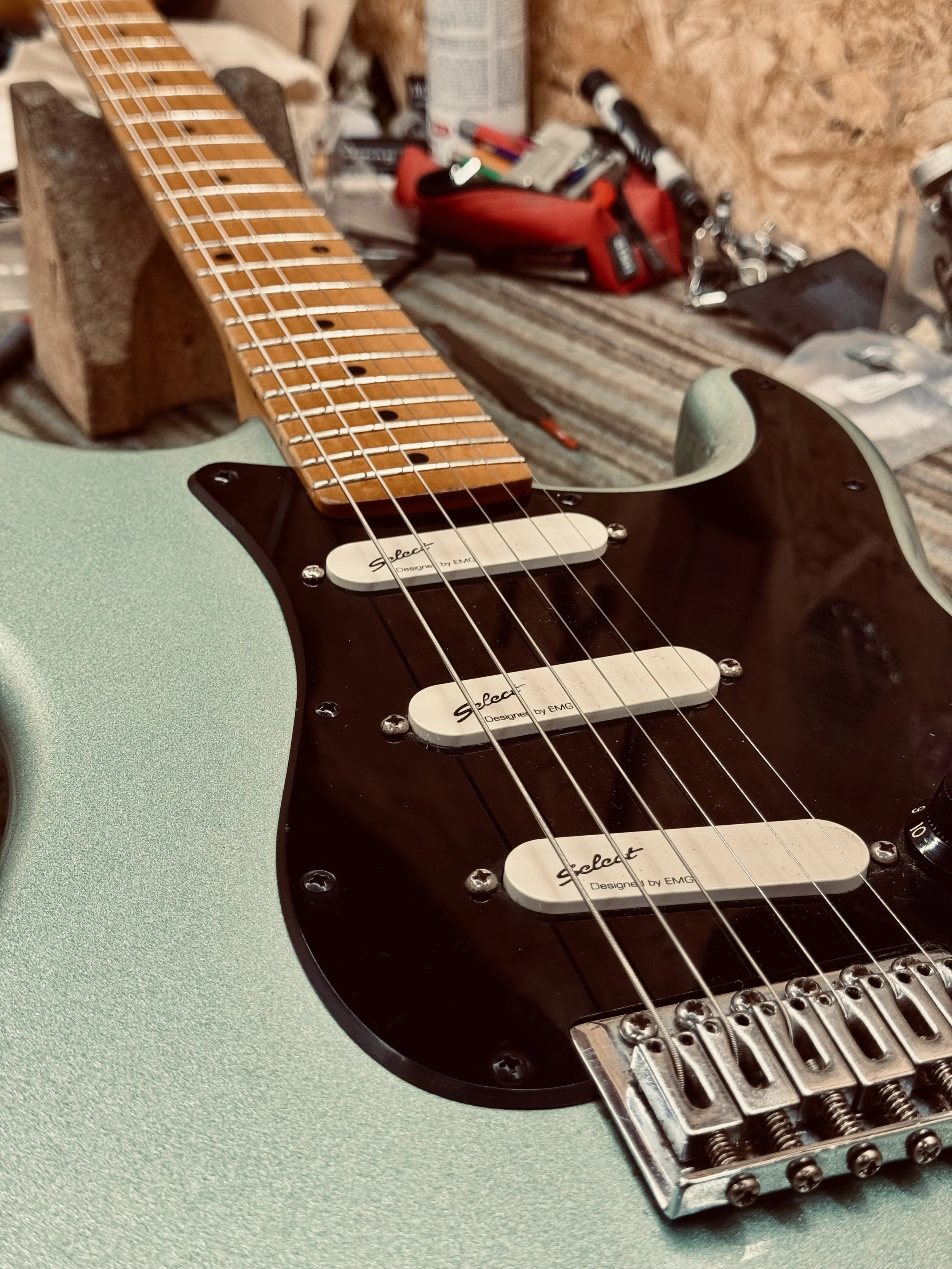 Close-up of a green electric guitar body with a black pickguard, three pickups, and a maple fretboard, in a workshop setting.