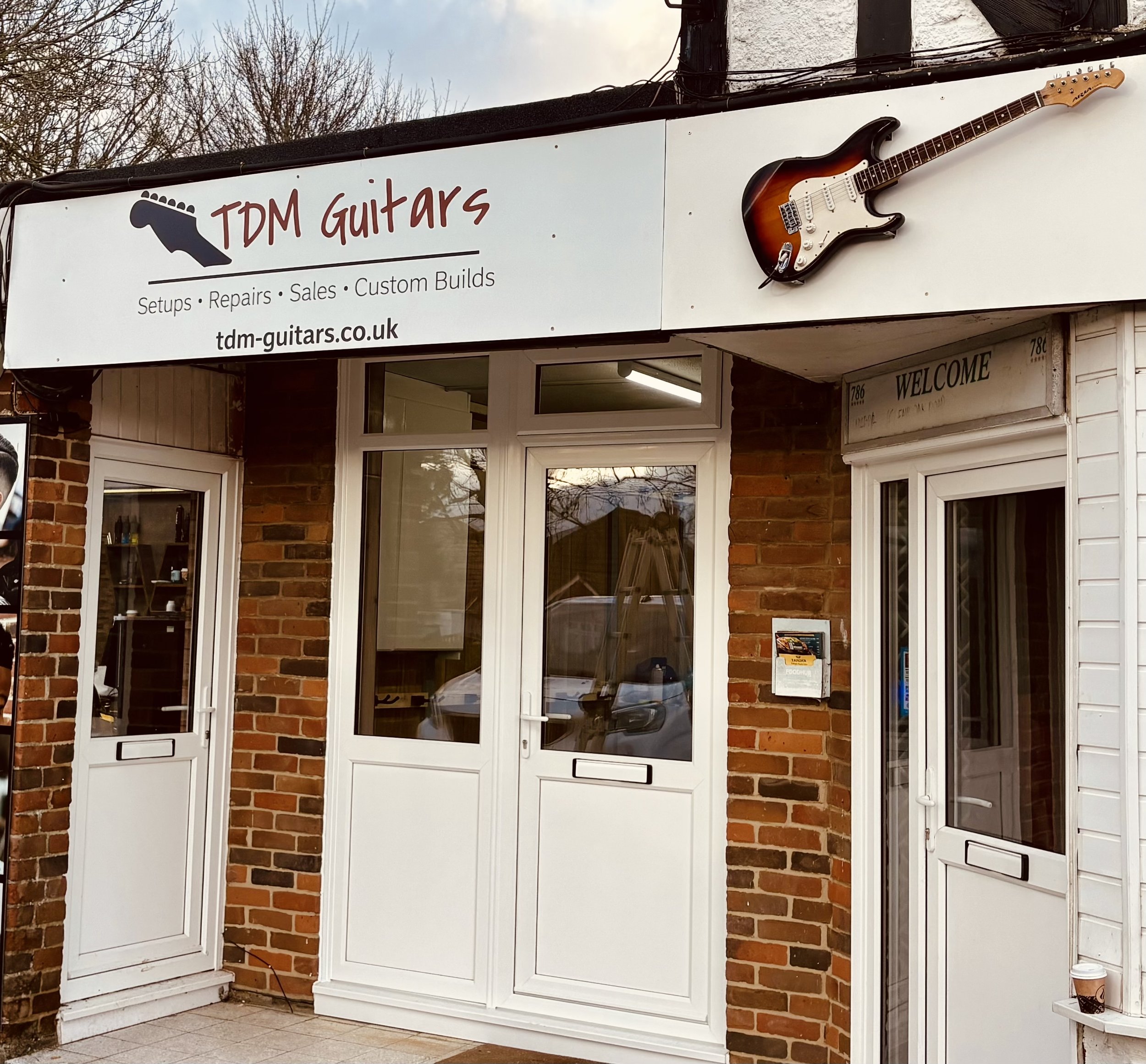 The storefront of TDM Guitars featuring a sign with a guitar graphic and the business name, along with an electric guitar mounted above the sign. The shop has a brick facade with two white doors and windows.