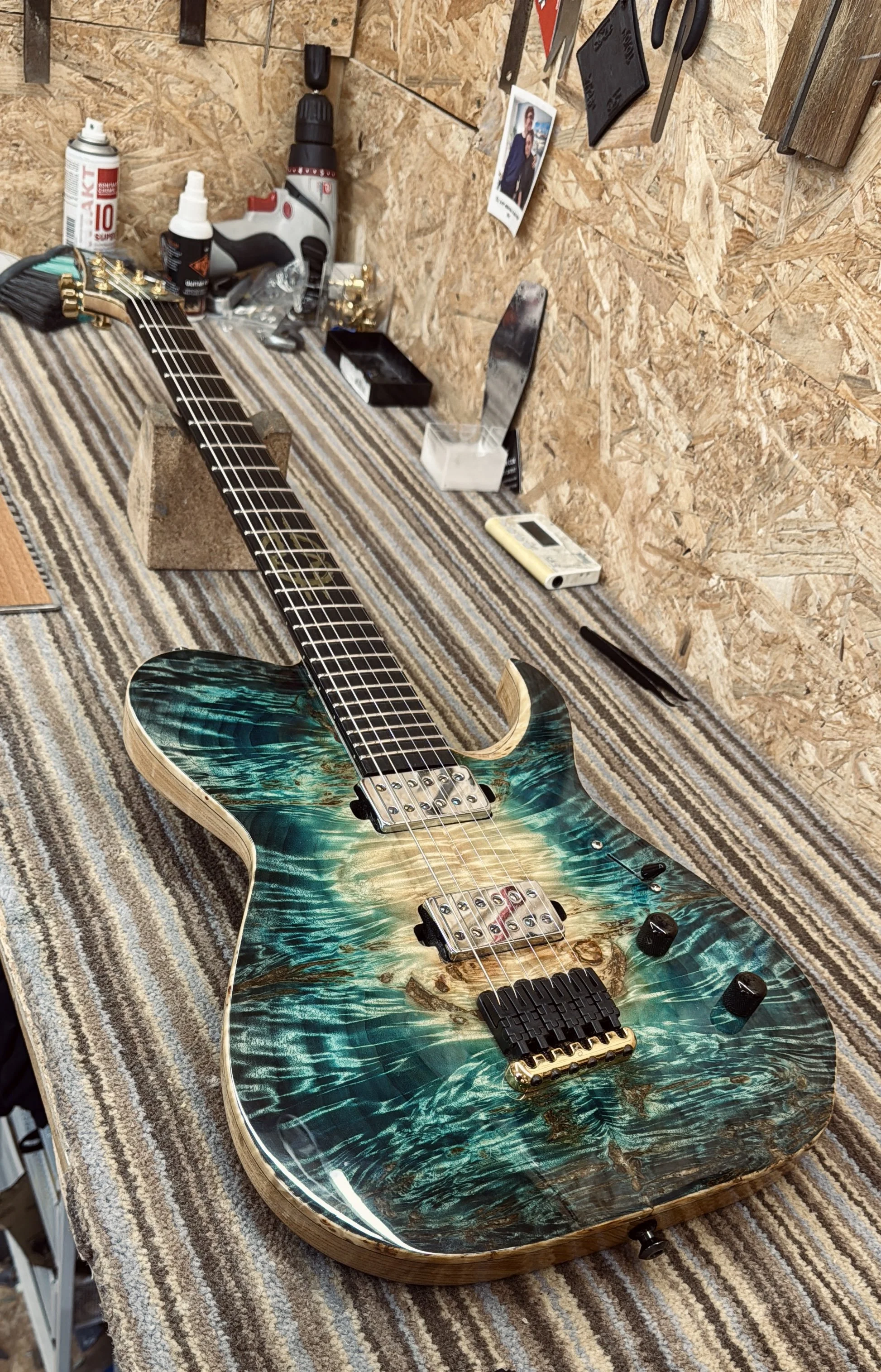A custom electric guitar with a swirling green, blue, and wood grain finish, placed on a striped workbench in a workshop. The guitar has a double humbucker pickup configuration, a black bridge, and black control knobs.