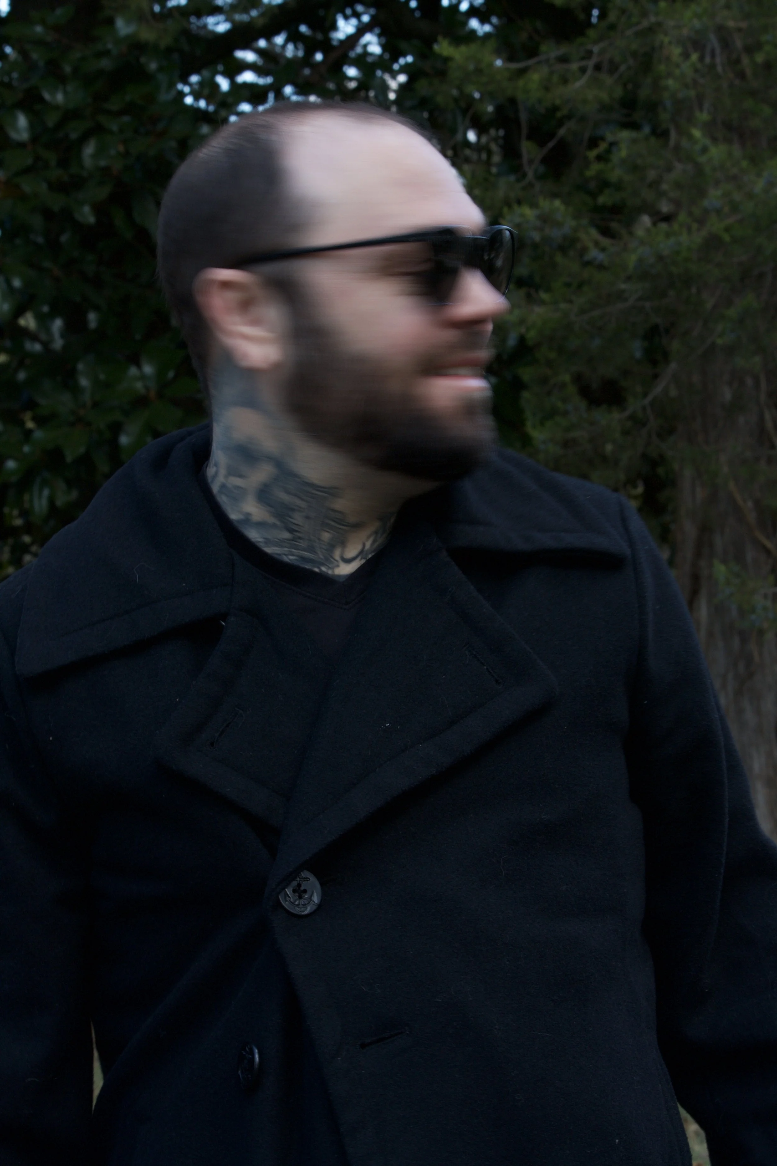 Blurry photo of a man with tattoos, black coat, sunglasses, with trees in the background.