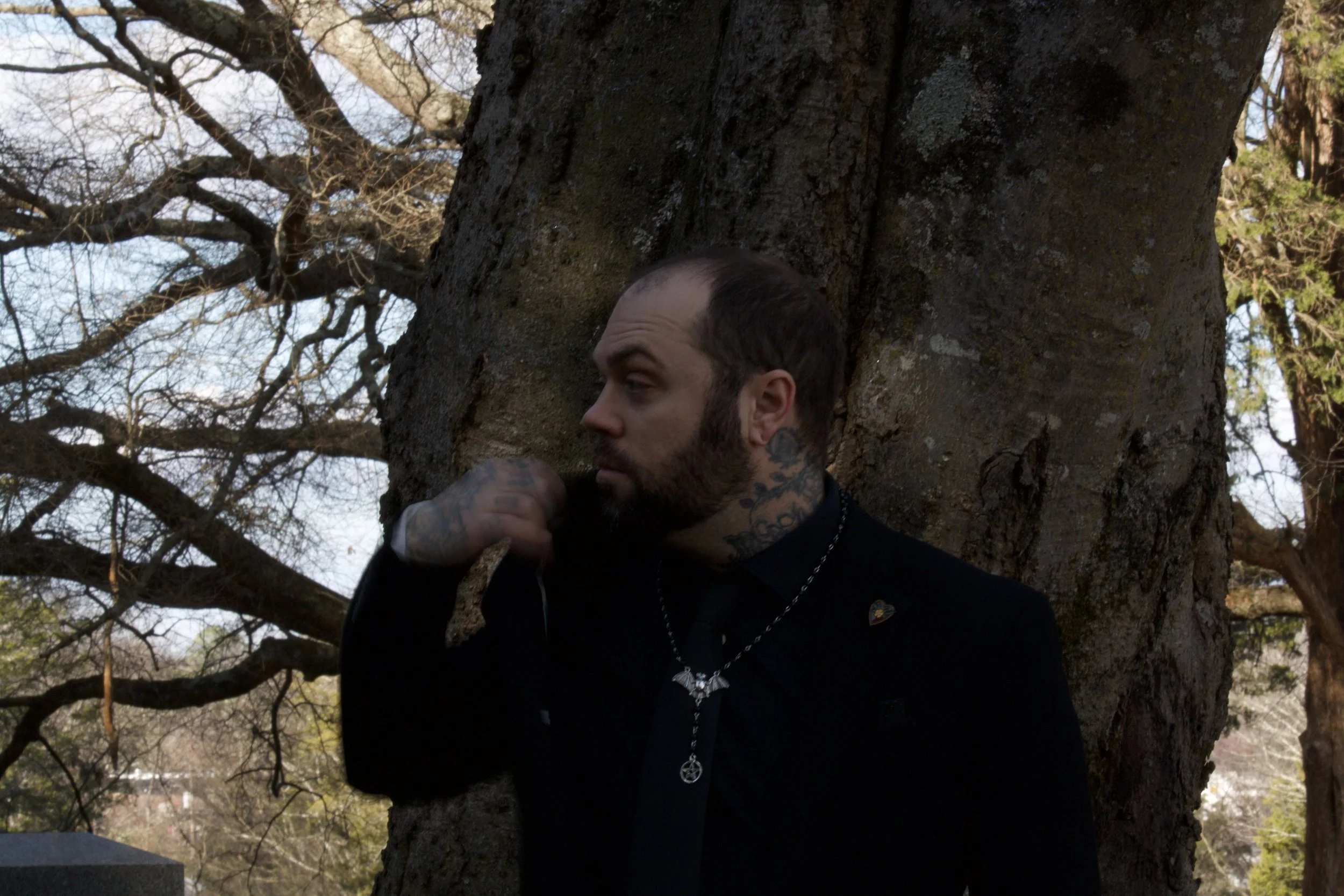 A man with tattoos and a beard leaning against a large tree, with leafless branches and some greenery in the background.