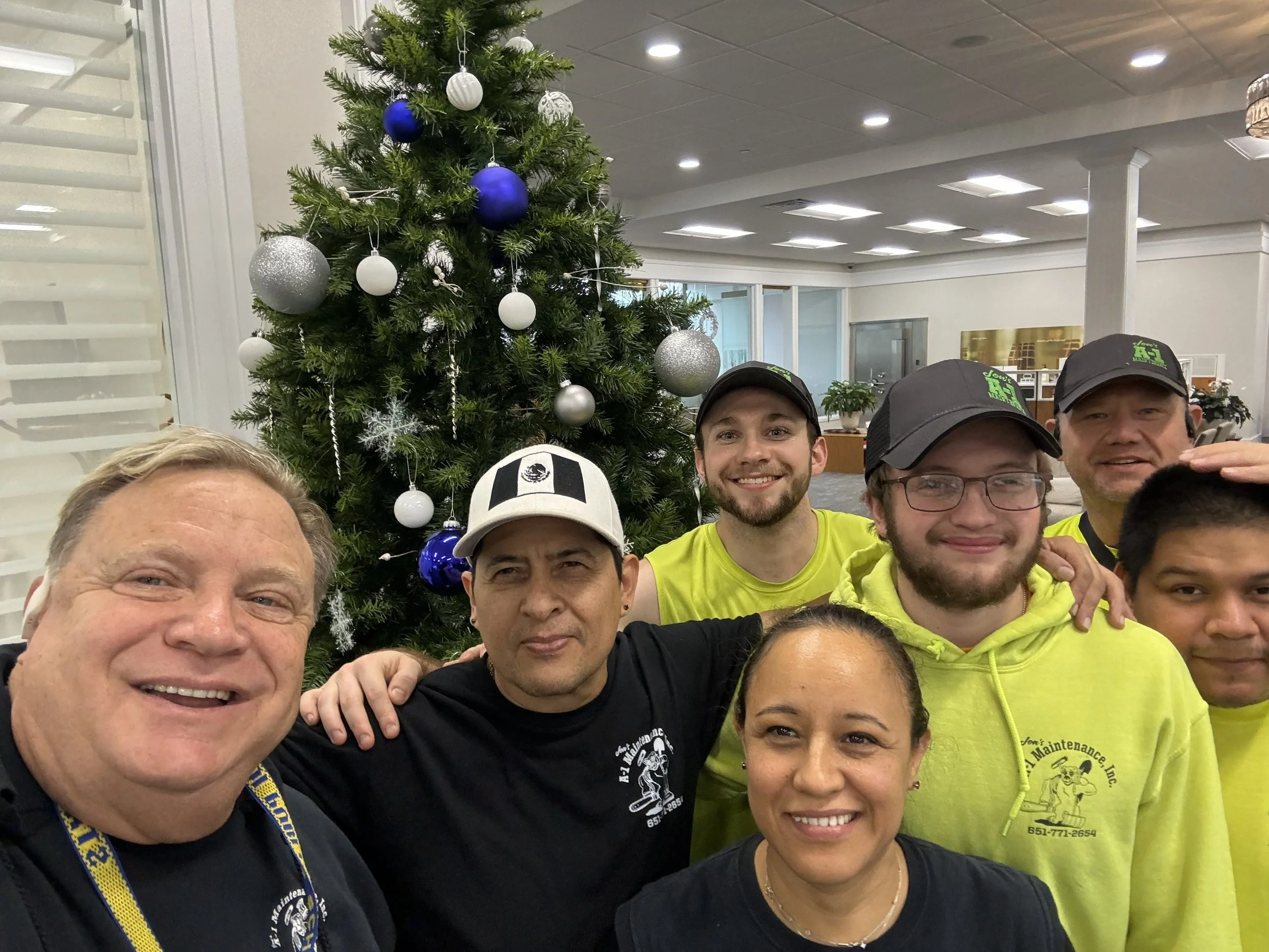 Group of people taking a selfie in front of a decorated Christmas tree inside an office or community center.