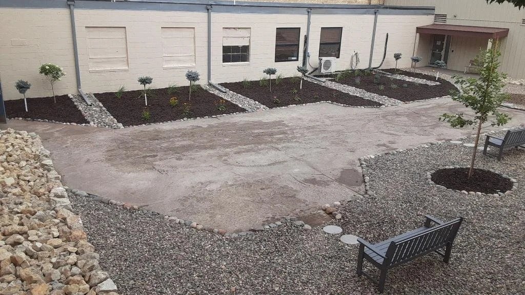 Backyard with landscaped garden beds, small trees, benches, and a concrete patio area.