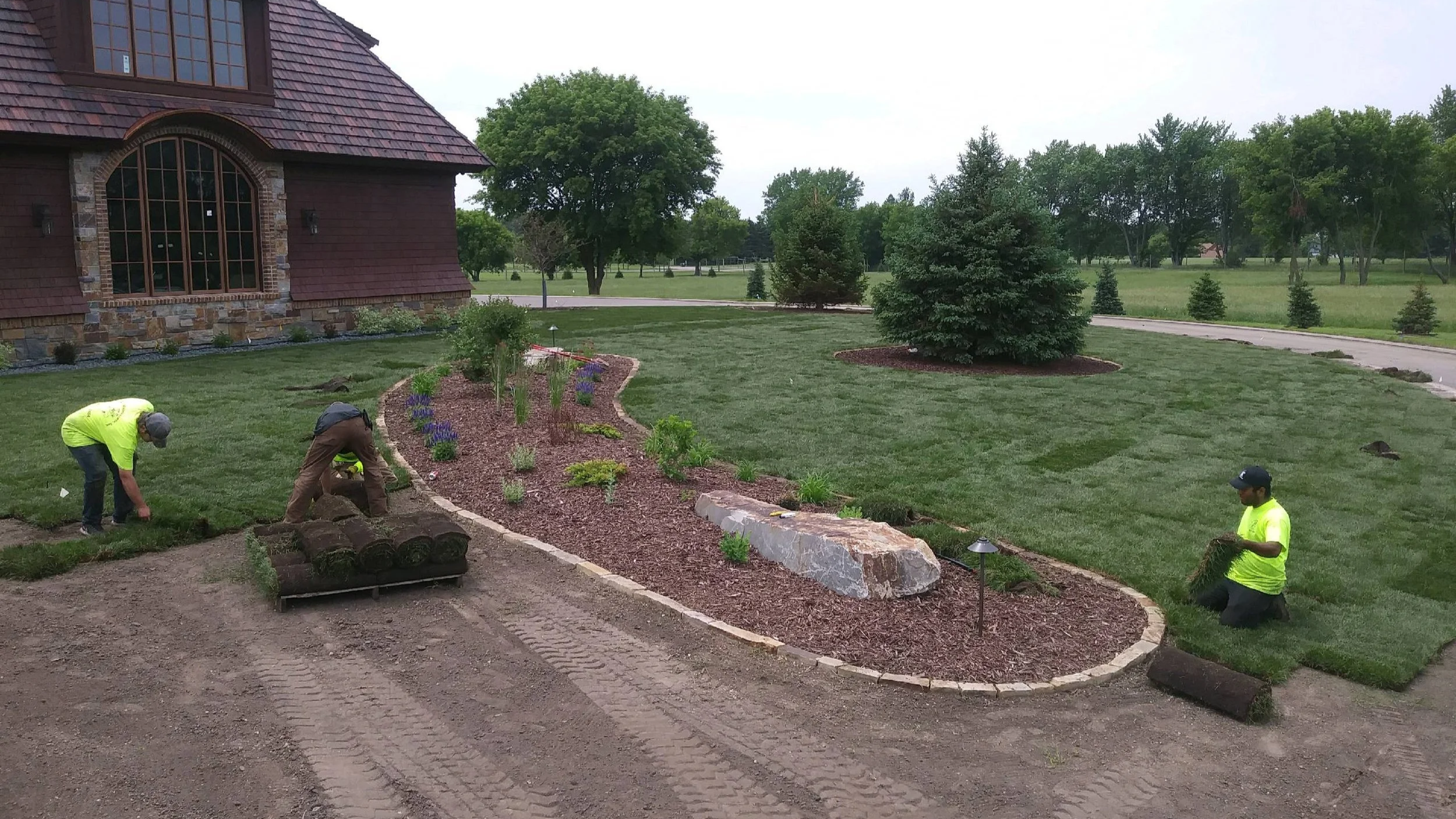 Three workers planting grass and landscaping a garden at a large house with a stone and wood exterior, surrounded by trimmed lawn and trees.