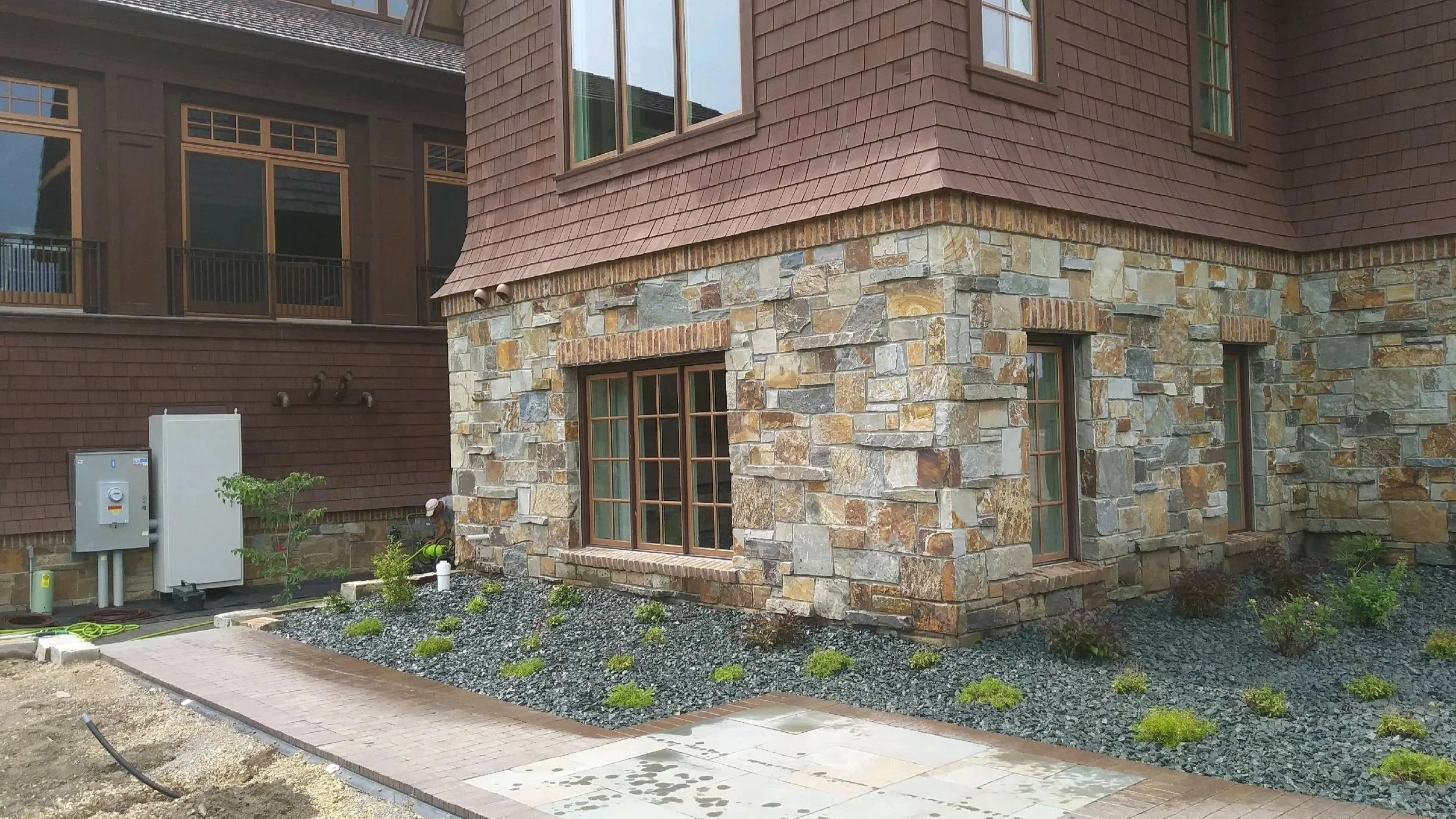 A residential house with a stone and wood exterior, multiple windows, and a landscaped front yard with small plants and gravel.