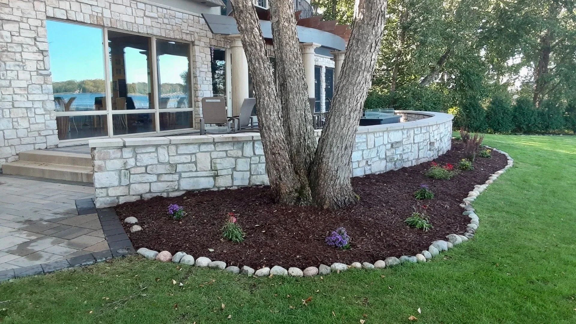 A landscaped backyard with a stone patio, a curved stone retaining wall, and a flower bed with small plants and purple flowers. A large tree is in the center of the flower bed, and there are outdoor chairs on the patio near a house with large glass w