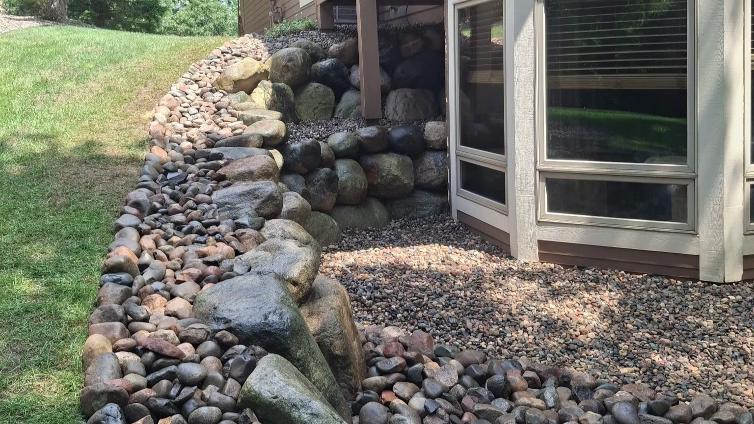 Decorative rock garden with large and small rocks along the side of a house, next to a grassy yard.