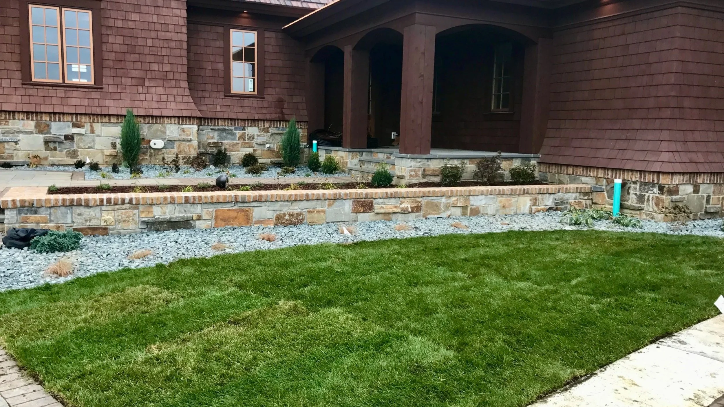 Newly landscaped front yard with green lawns, a rock-bordered flower bed, small shrubs, and young trees in front of a brick house with dark brown siding and windows.