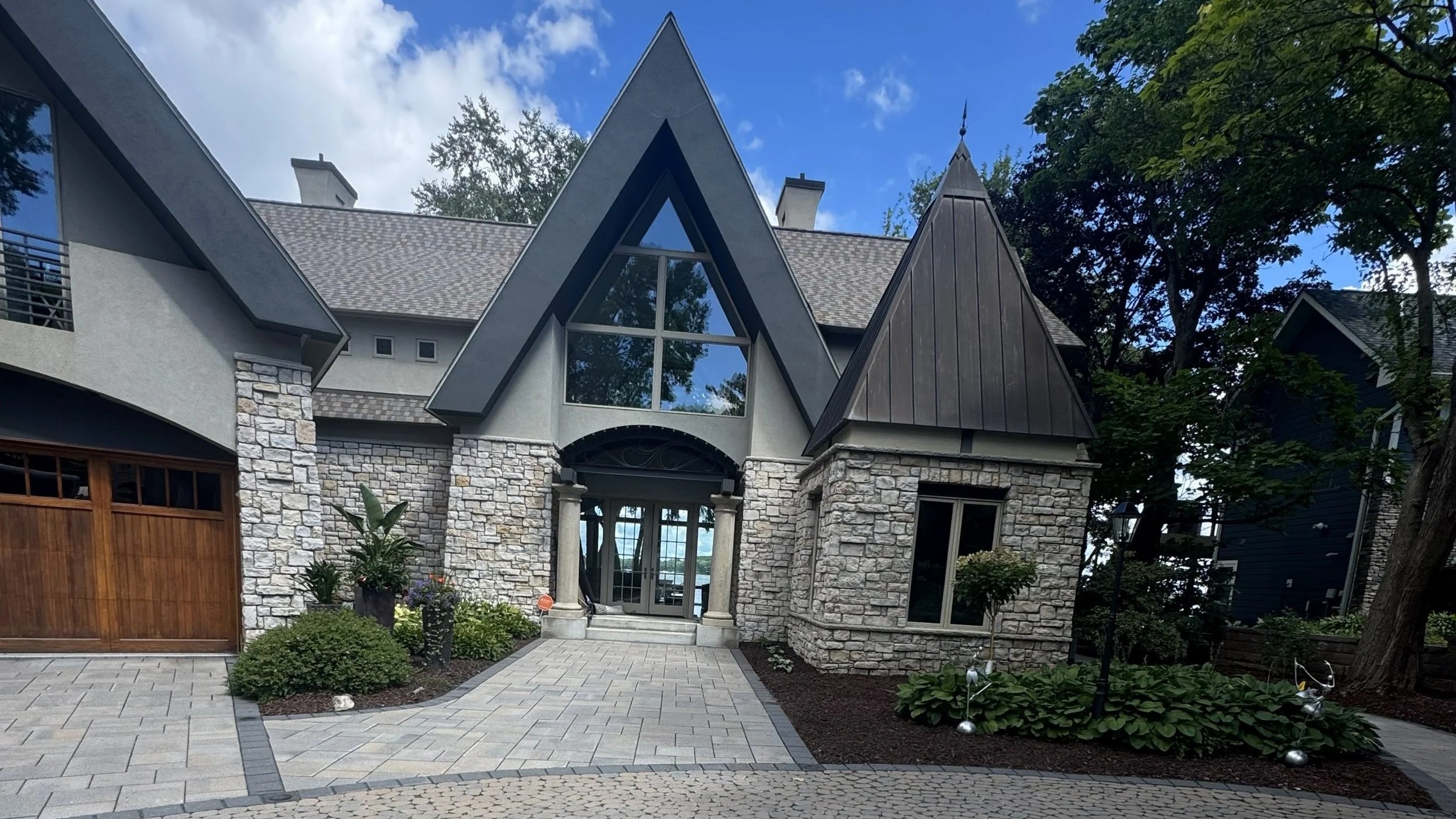 Exterior view of a modern house with stone and wood accents, large triangular window, surrounding landscaped garden.