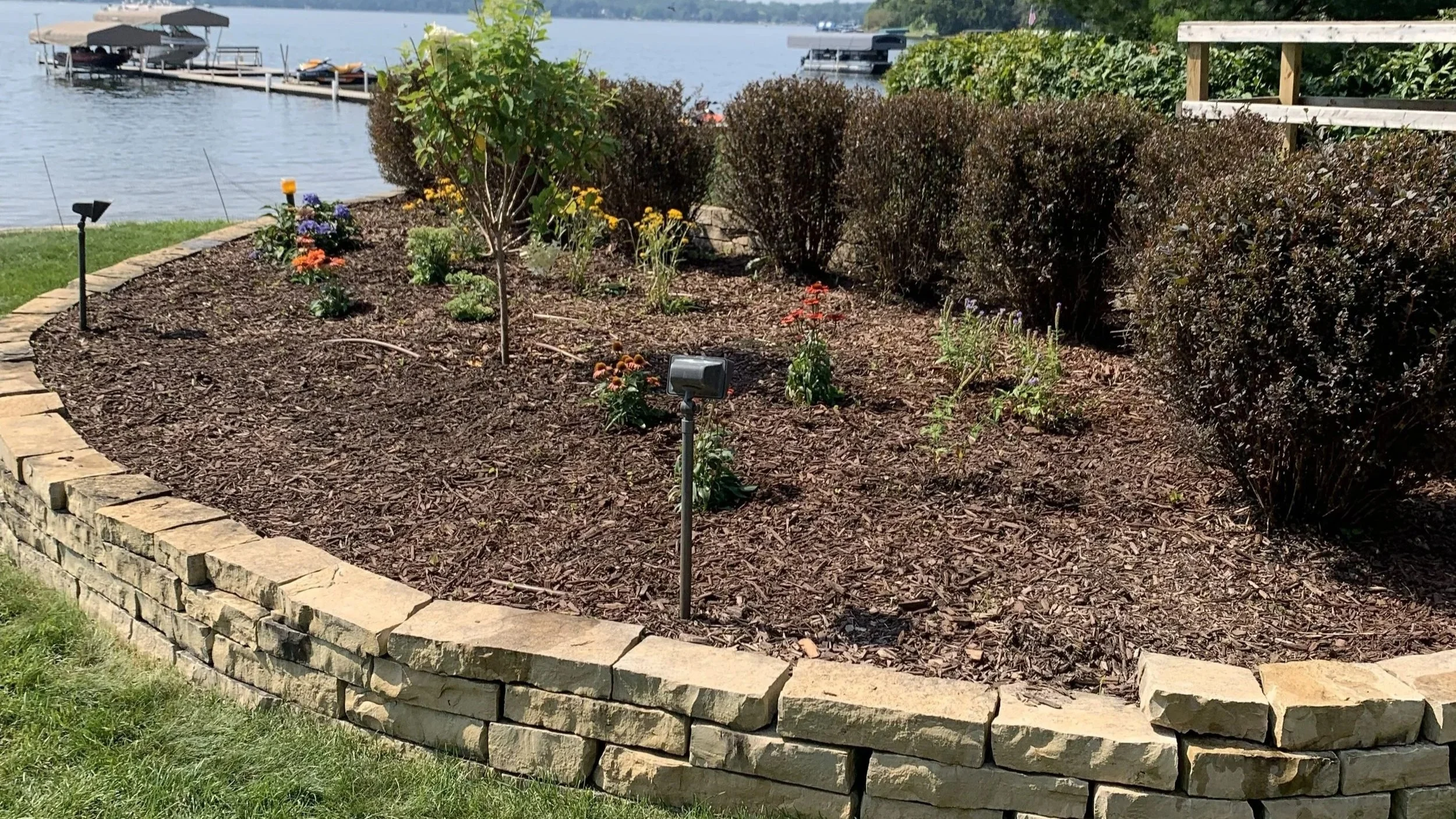 A landscaped garden bed with a stone border, various plants, bushes, and flowers, with a body of water and boats in the background.