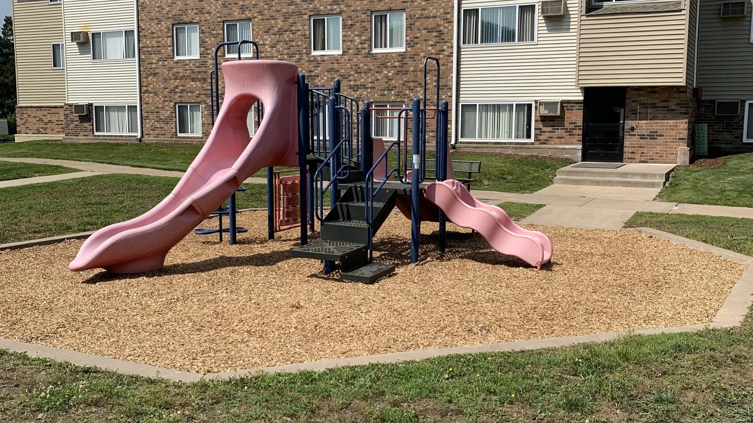 A playground with pink slides, black steps, and blue railings located in front of an apartment building. The ground is covered with wood chips, and there are sidewalks and grass surrounding the area.