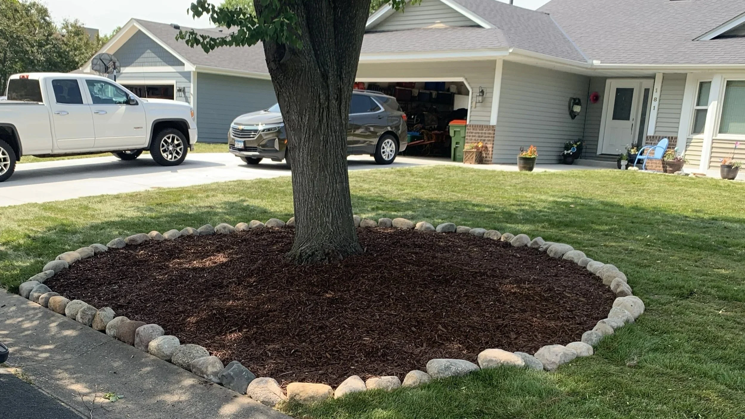 Residential front yard with a tree surrounded by a border of rocks and mulch, adjacent to a driveway with parked cars, and a house with a porch and decorative outdoor items.