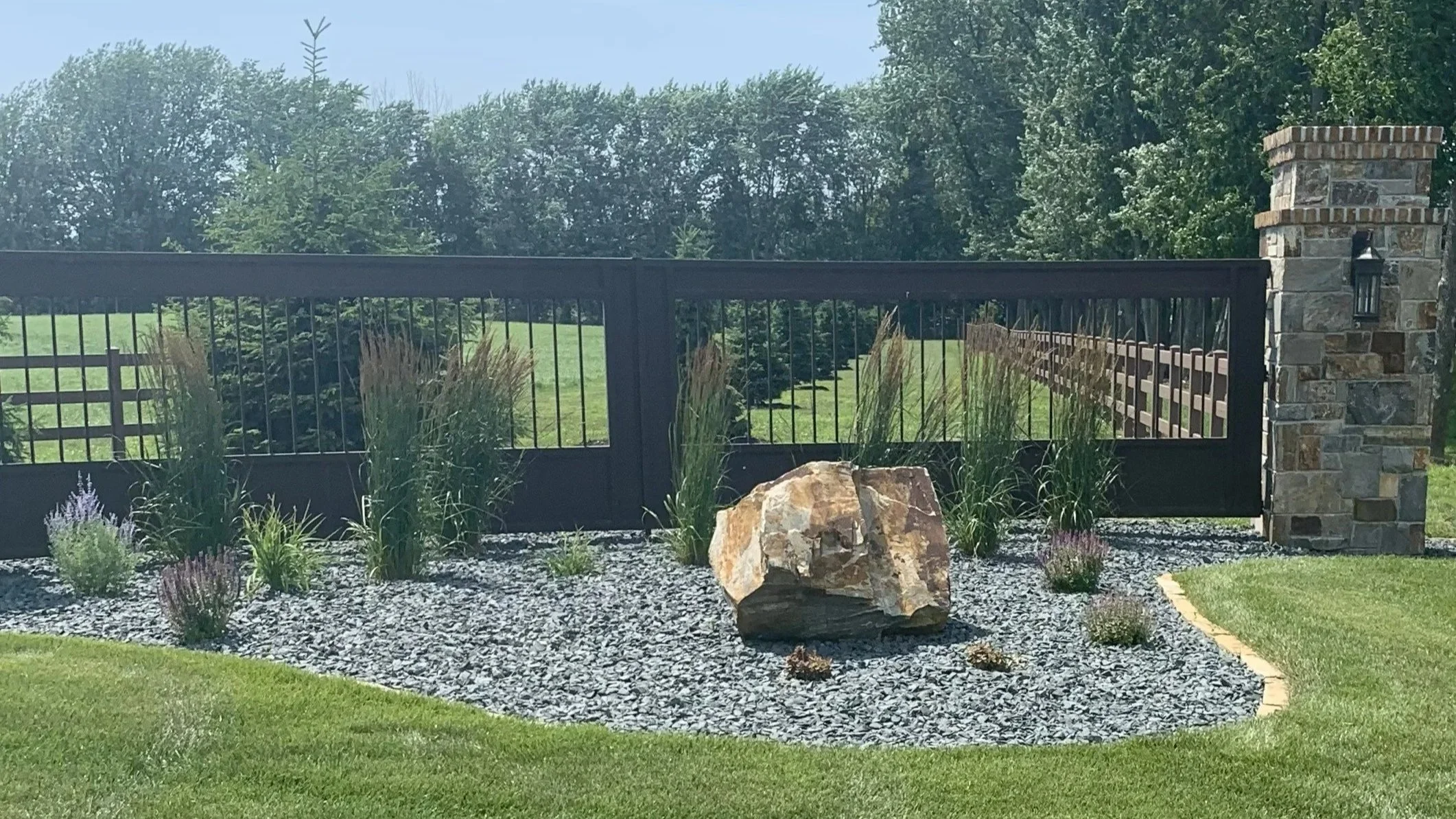 A landscaped yard features a gravel bed with ornamental grasses, a large rock, and smaller plants, enclosed by a black metal gate and stone pillars, with a background of green trees and rolling field.