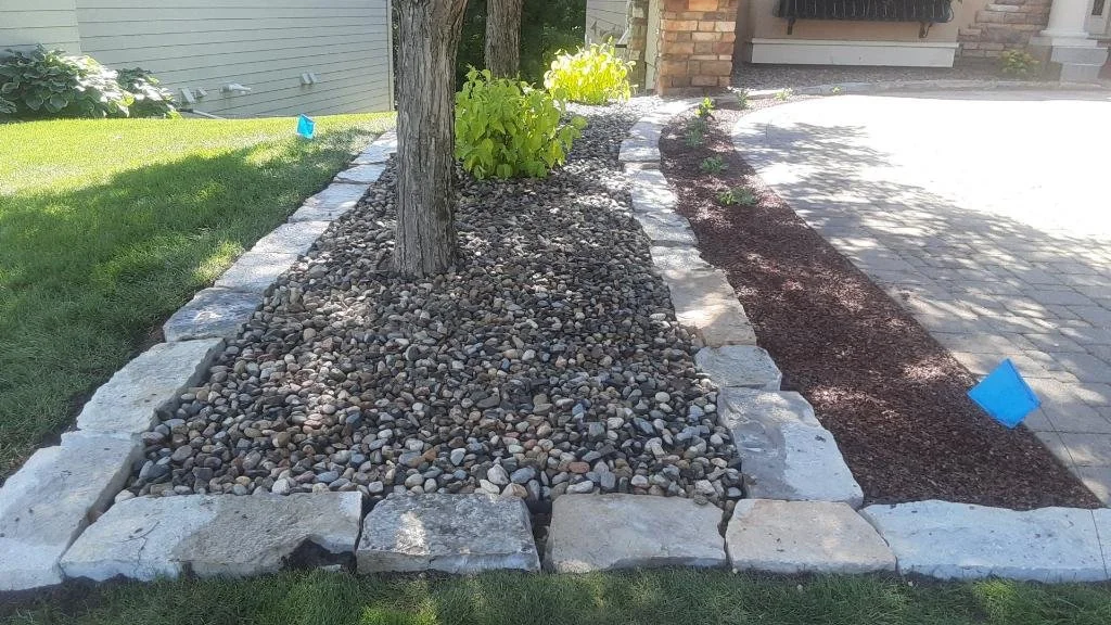 A landscaped garden bed with a small tree, green plants, and decorative rocks, bordered by large gray stone bricks, with newly added soil and mulch on a paved driveway.