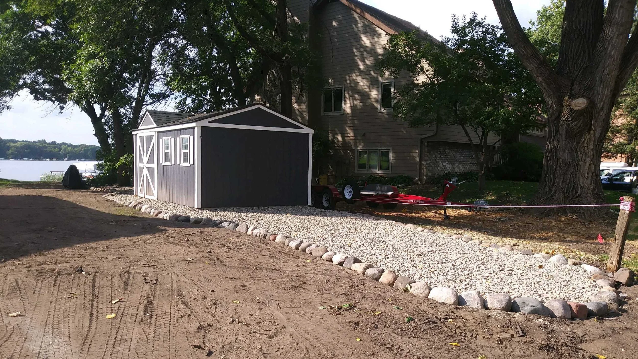 A small gray shed with white trim adjacent to a large tree, with a gravel pathway in front, near a lake and residential buildings in the background.