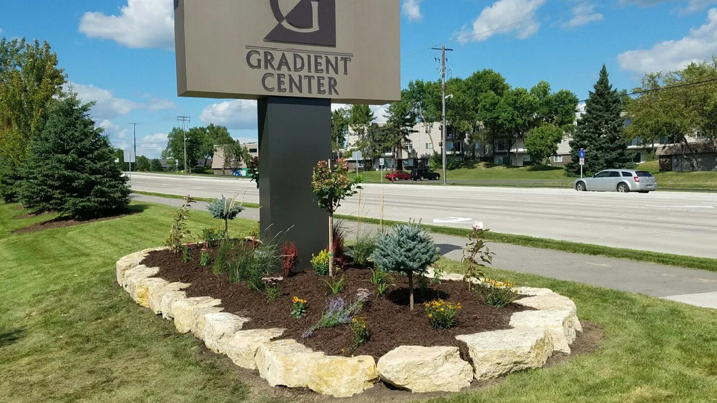 A landscaped area with a stone border, small trees, flowering plants, and mulch, in front of a sign that reads 'Gradient Center' beside a street with cars and residential buildings in the background.