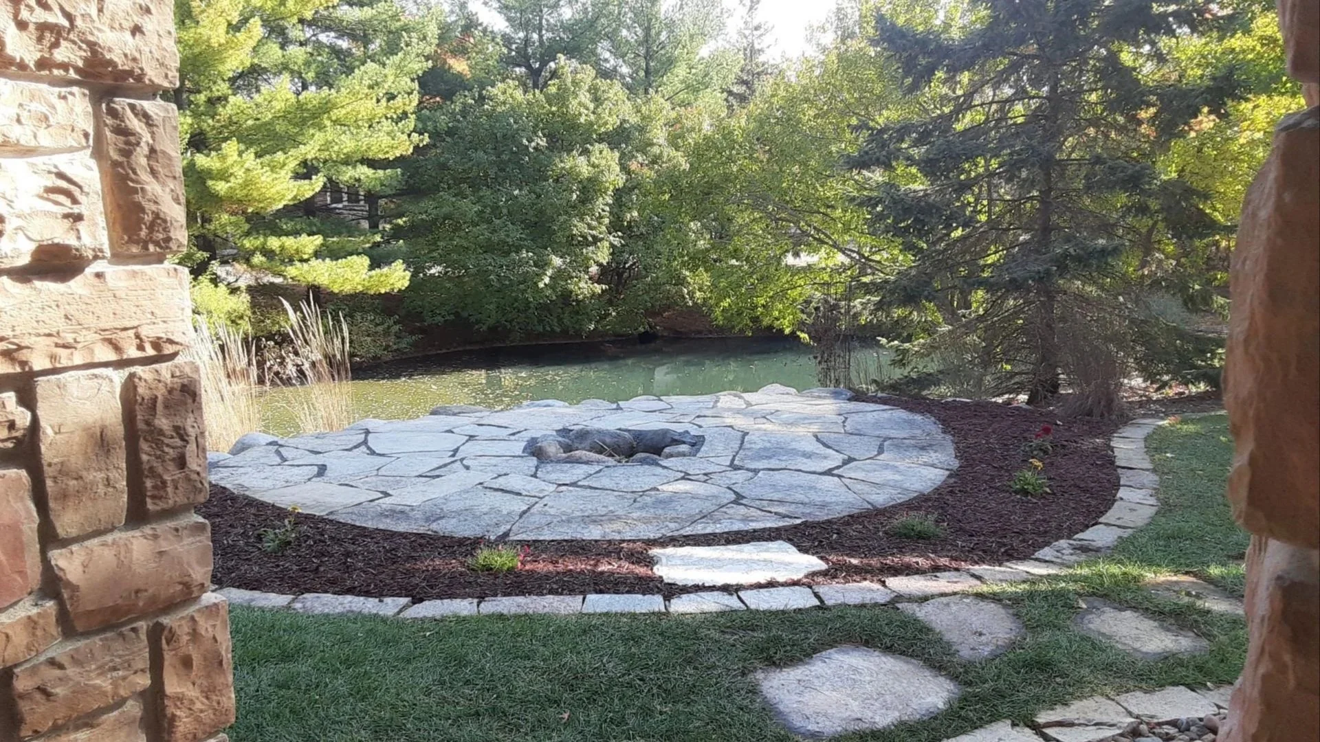 View through a stone archway onto a landscaped backyard with a small pond, stone patio, and surrounding trees.