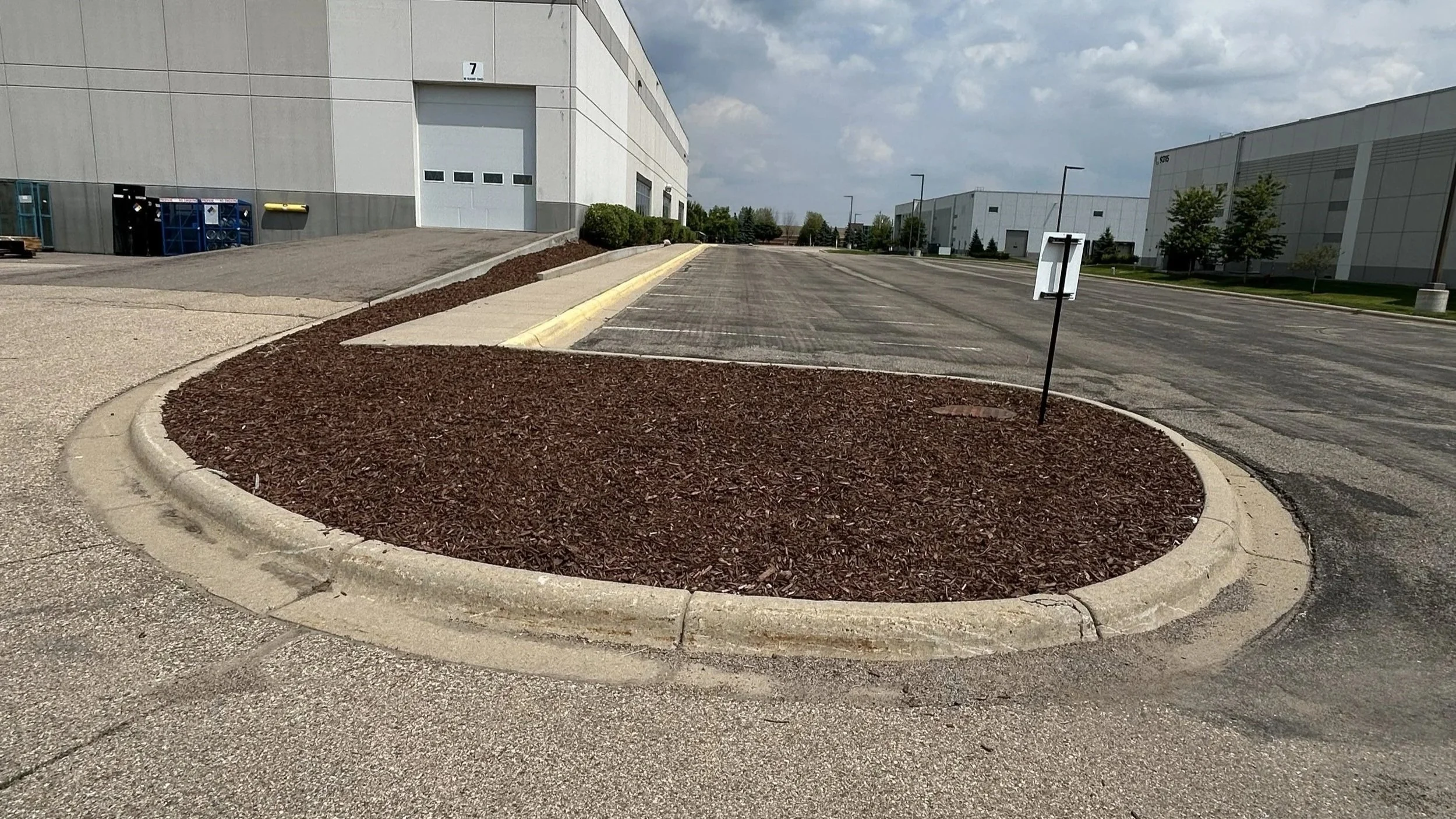 Empty parking lot with a landscaped island in the foreground, surrounded by industrial buildings, under a cloudy sky.