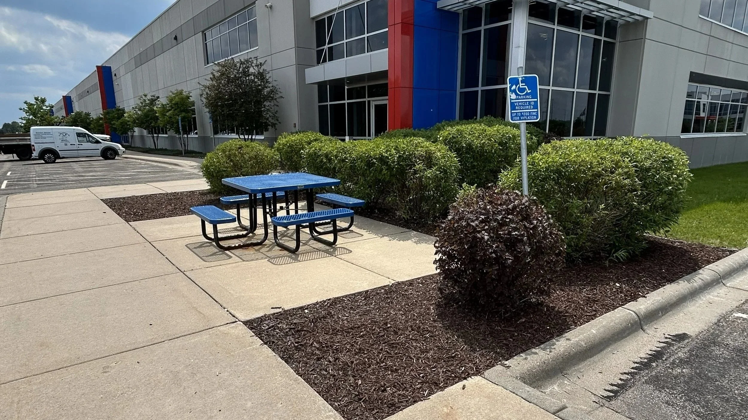 A handicapped parking sign near a sidewalk with bushes, and a blue metal picnic table in front of a modern building with large windows.
