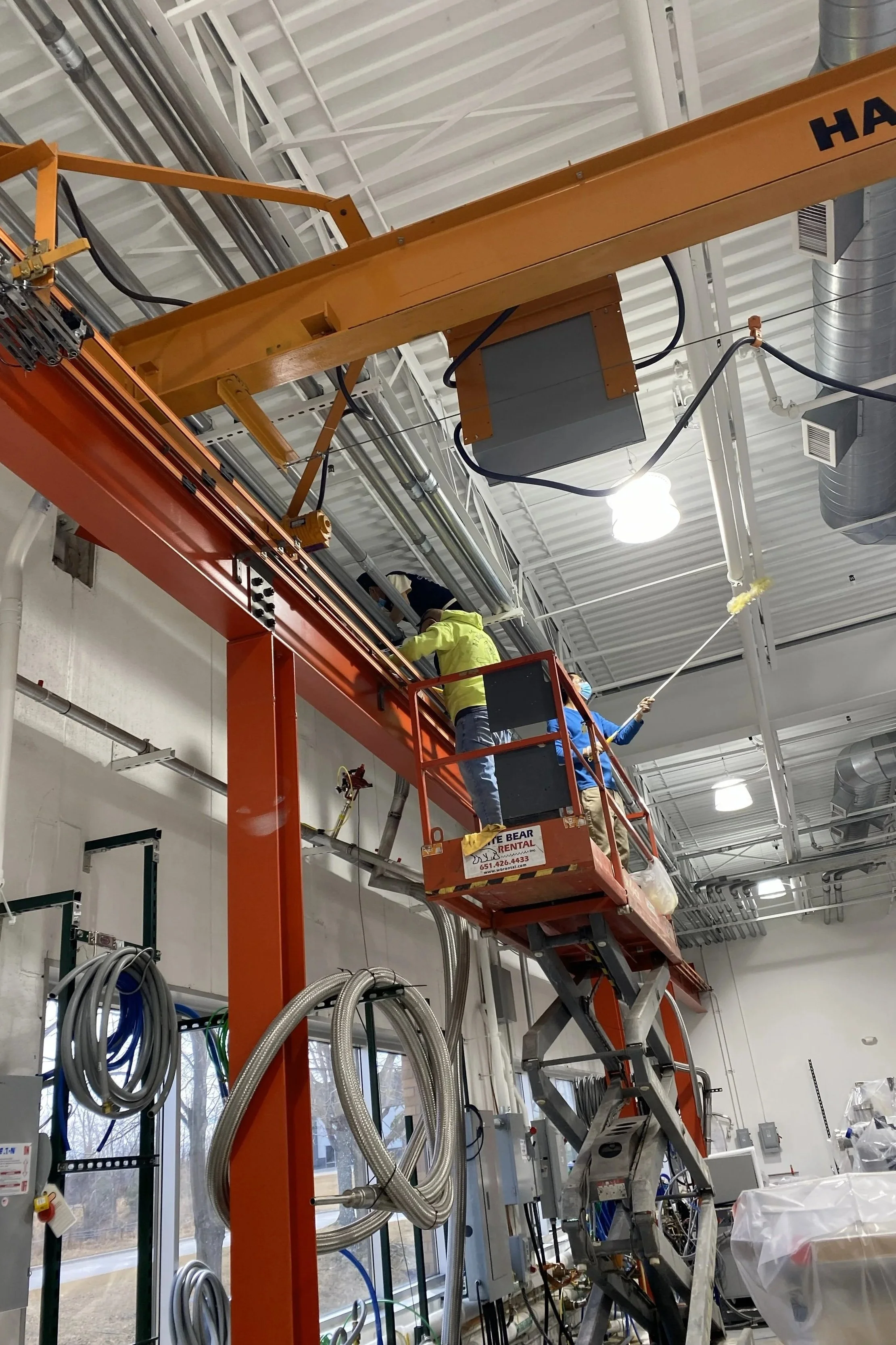 Workers on an orange scissor lift working on ceiling piping and electrical systems inside an industrial or commercial building.