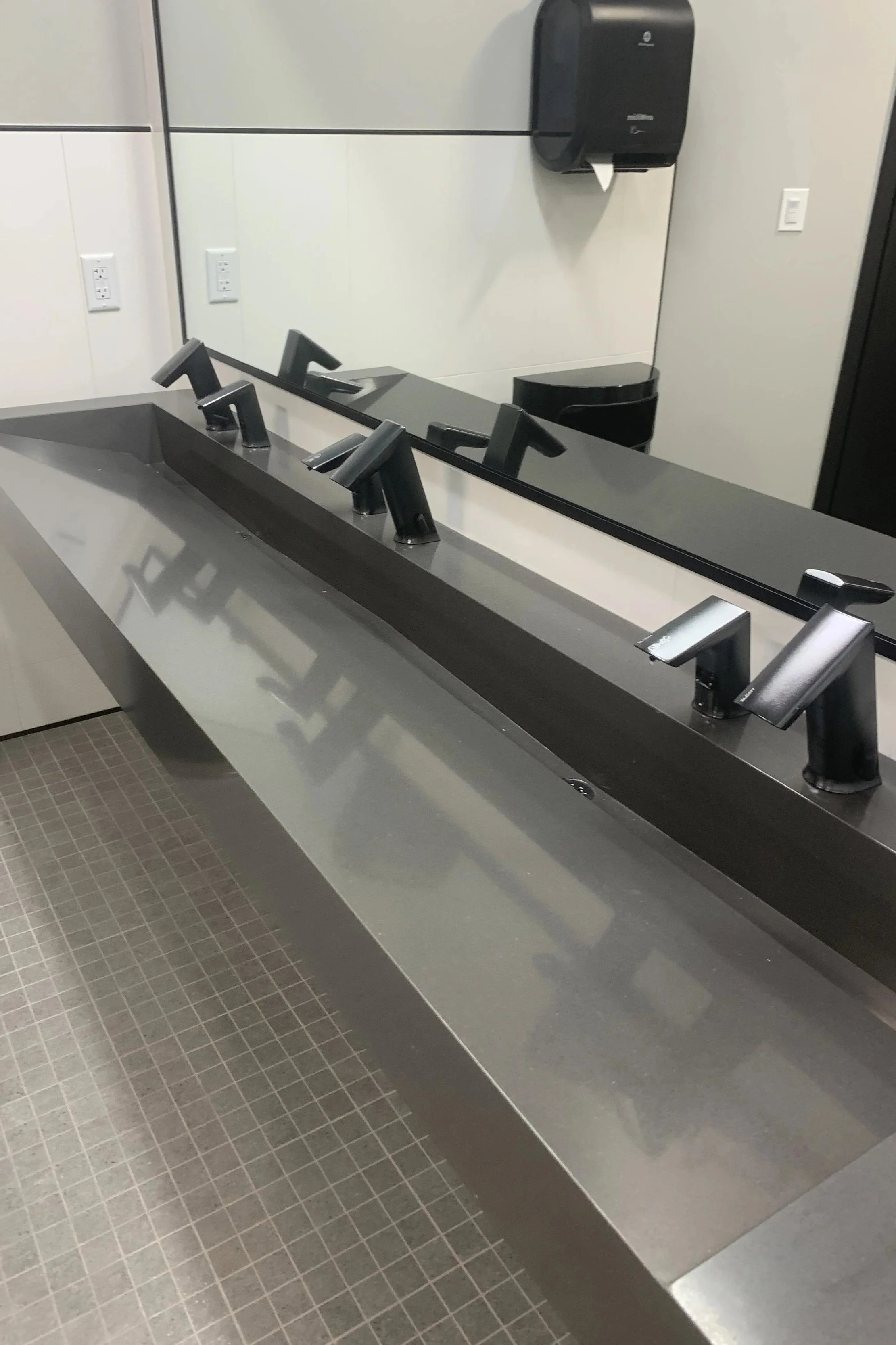 A modern bathroom countertop with three black faucets, a large mirror above, a paper towel dispenser on the wall, and a trash can in the background.