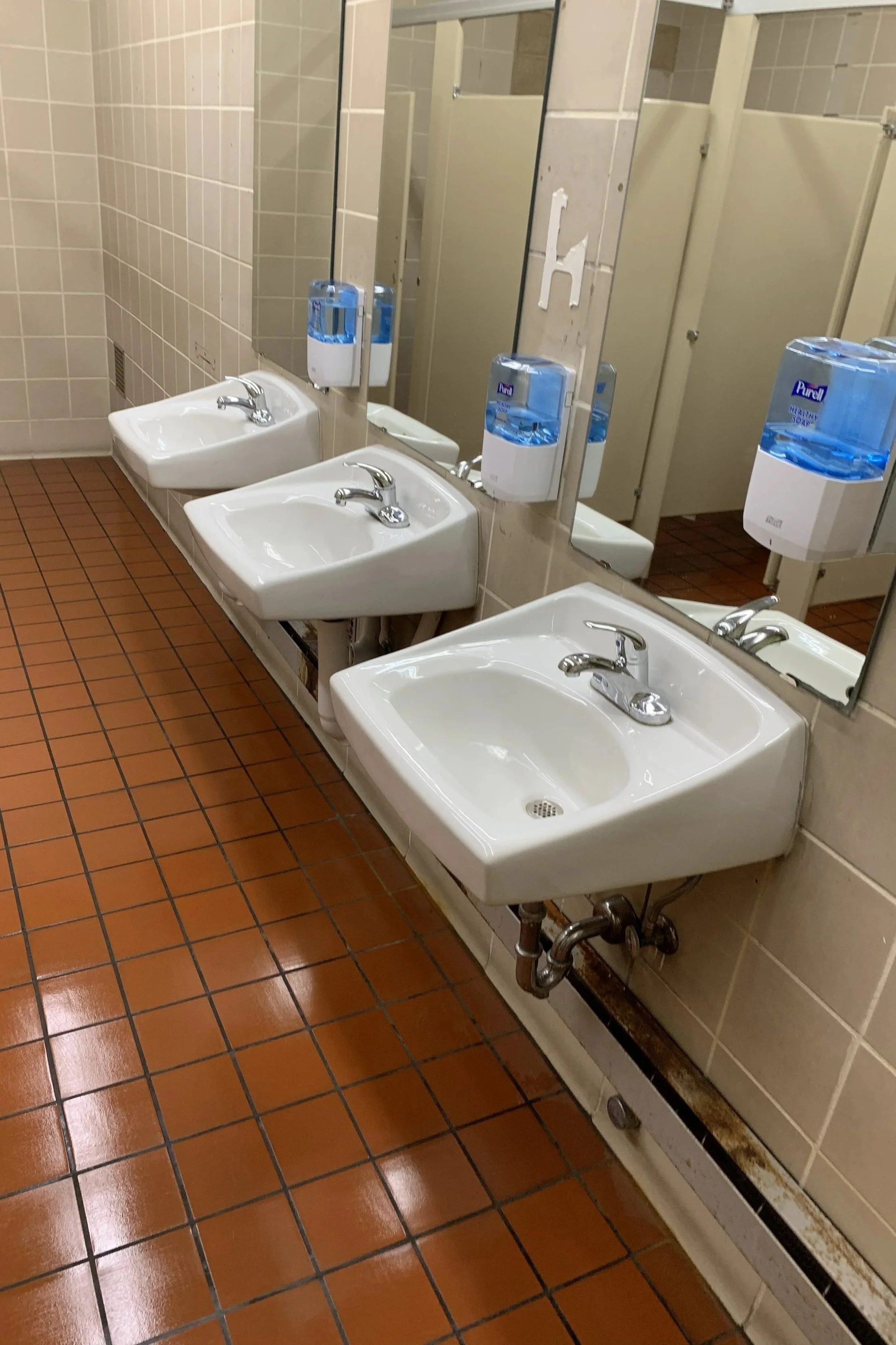 Three white sinks with individual faucets are mounted on the wall in a restroom, with a large mirror above them. There are two blue soap dispensers attached to the wall between the sinks and the mirror. The restroom has a brown tiled floor, cream til