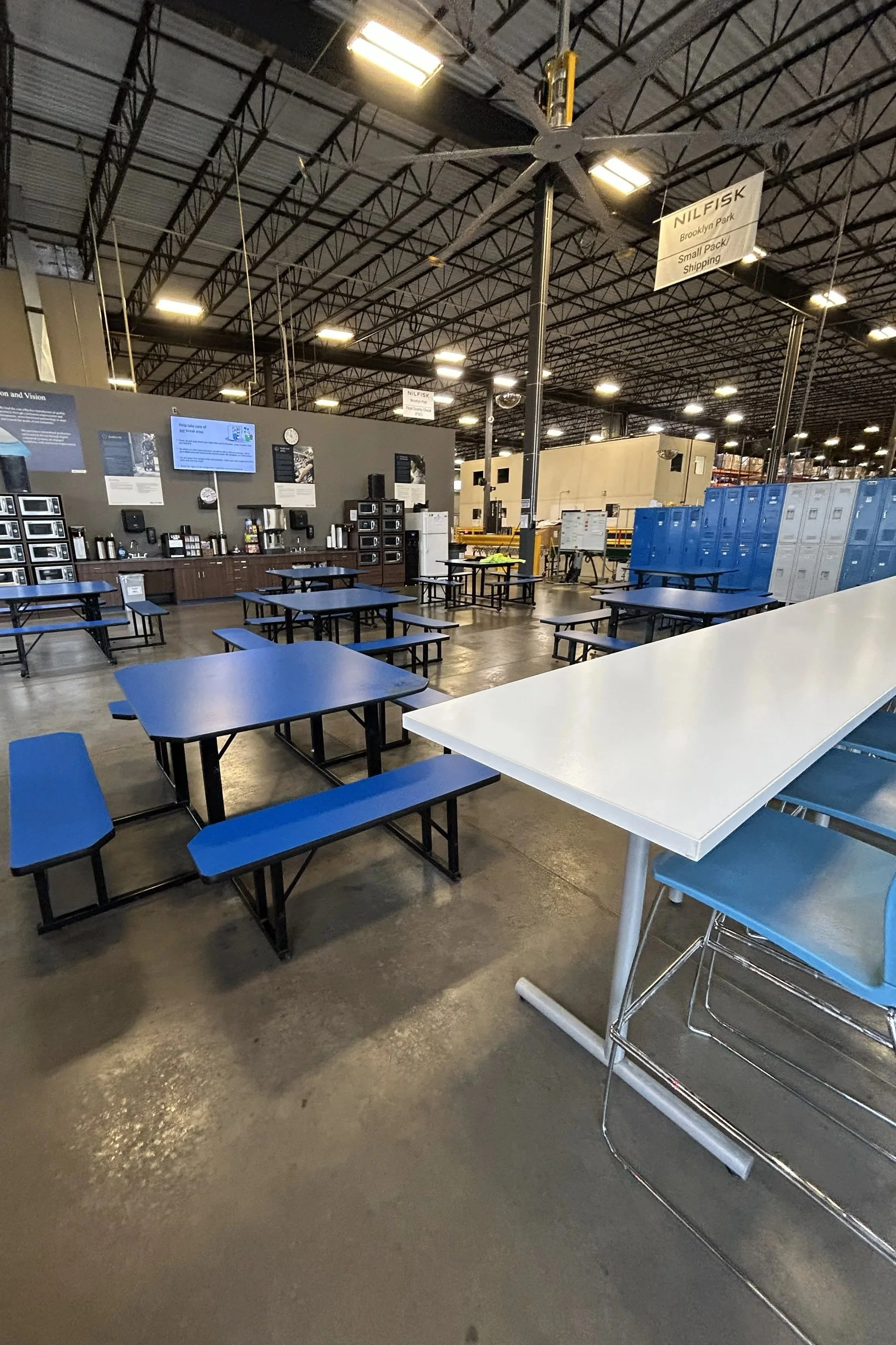 Empty cafeteria or break room area in a warehouse or industrial facility with blue benches and tables, lockers, a long white table, and wall-mounted monitors.